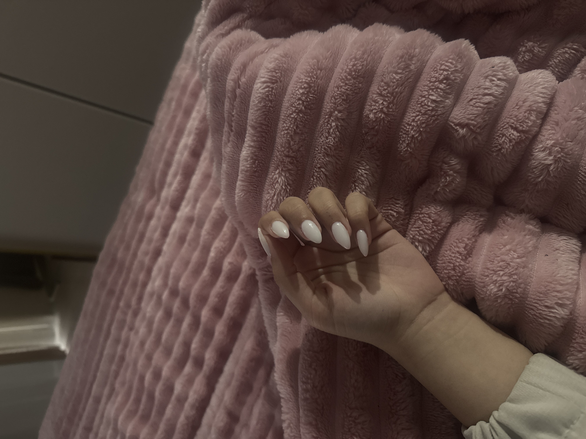 The photograph depicts a hand resting on a pink, plush, horizontally ribbed blanket. The hand has pale skin and features manicured nails painted a solid white color, shaped to a rounded point. The wrist of the hand is visible, extending into a white sleeve. In the background, to the left, is a blurred section of what appears to be a light-colored drawer or piece of furniture. The location appears to be indoors. No city is discernable.