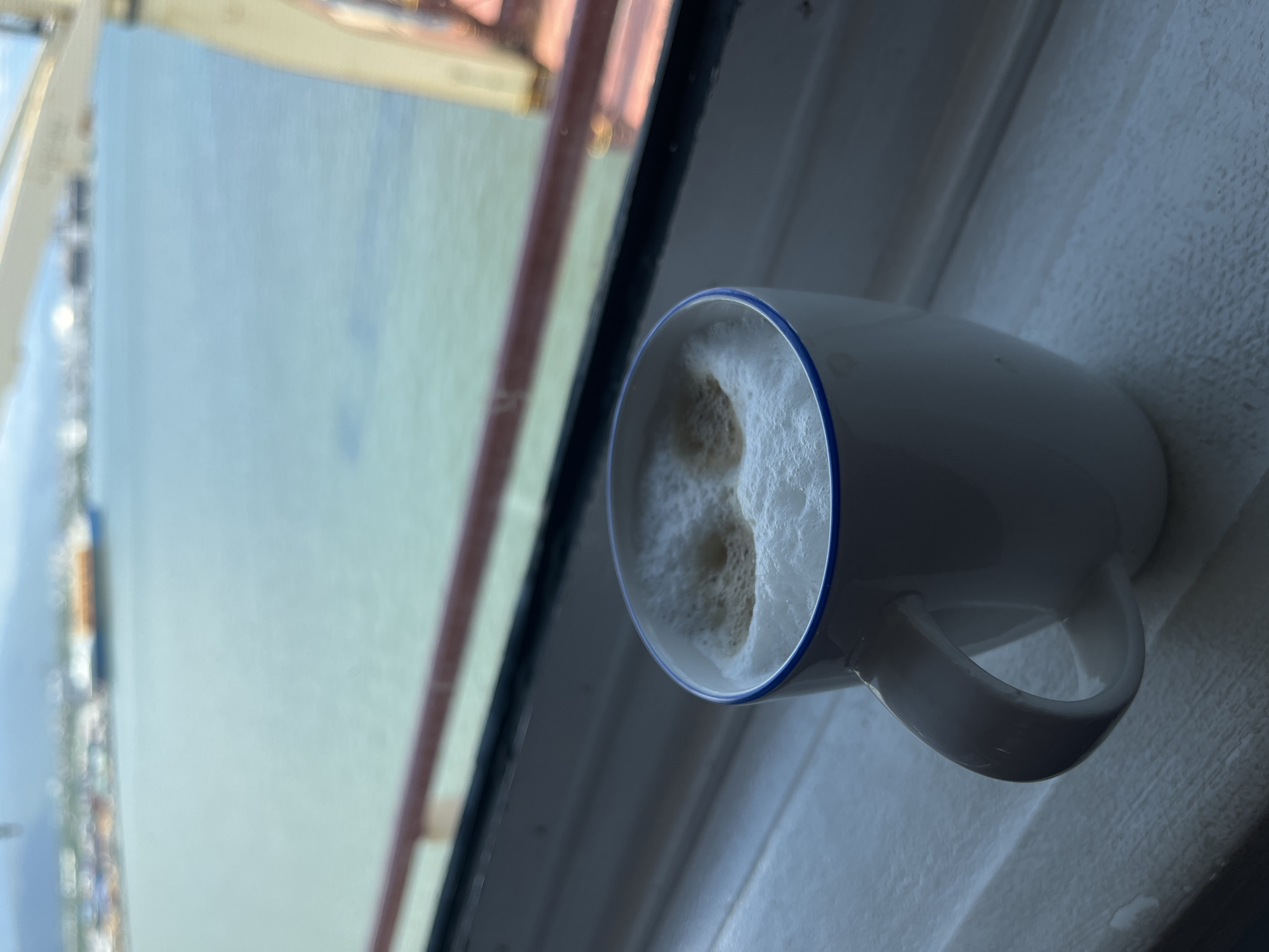 A mug of beverage with white foam sits on a windowsill. The mug is white with a blue rim and a handle. Outside the window, a body of water is visible. Along the water's edge, there is a coastline.
