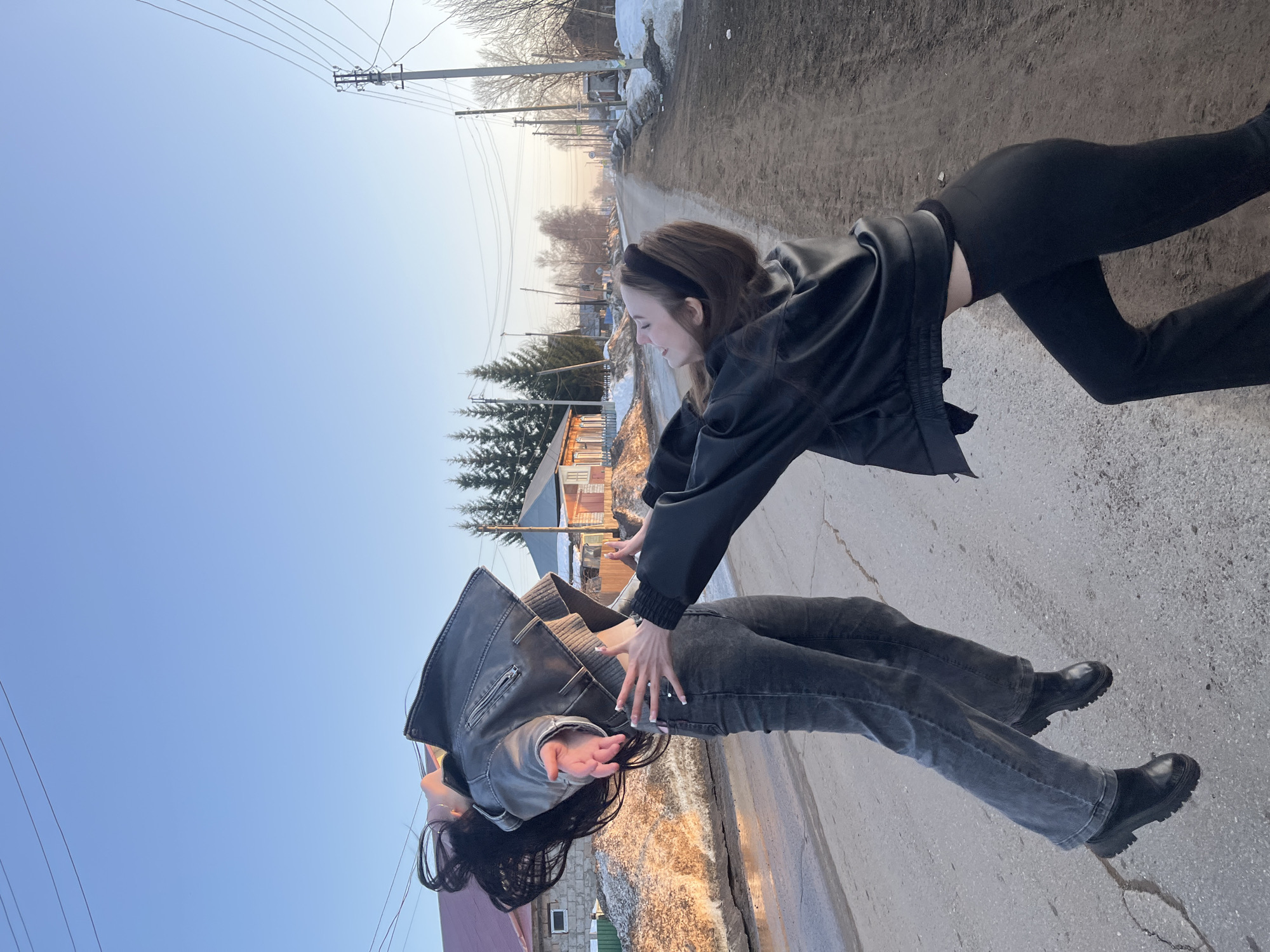 This image captures two young adult females interacting on a semi-paved road in the city of Фоки, Russia. The scene is illuminated by the low, warm light of late afternoon or early evening, likely during late winter or early spring, as evidenced by melting snow and bare deciduous trees.
One female, positioned on the left side of the frame, is depicted mid-air, appearing to jump or float. She is oriented slightly to her right, with her head tilted back and long dark hair flowing upwards and backward. Her arms are extended forward with open palms. She is dressed in a dark gray or black cropped leather-look jacket, a light-colored crop top, dark gray wide-leg jeans, and black chunky-soled boots.
The second female, situated to the right, is crouched slightly, leaning forward with both arms extended and palms open towards the first individual. She wears a black leather-look bomber jacket, a black top revealing a portion of her midriff, and black flared trousers. Her brown hair is styled with a black headband, and her gaze is directed towards the airborne female.
The ground consists of a two-lane road with asphalt on the left and a dirt or gravel shoulder on the right, displaying cracks and uneven surfaces. Piles of dirty, melting snow and slush line both sides of the road, interspersed with puddles. The background features several detached, single-story houses with various roof colors, coniferous trees to the left, and bare deciduous trees further down the road. Multiple utility poles carrying power lines are visible along both sides of the road, extending into the distant horizon under a clear, light blue sky.