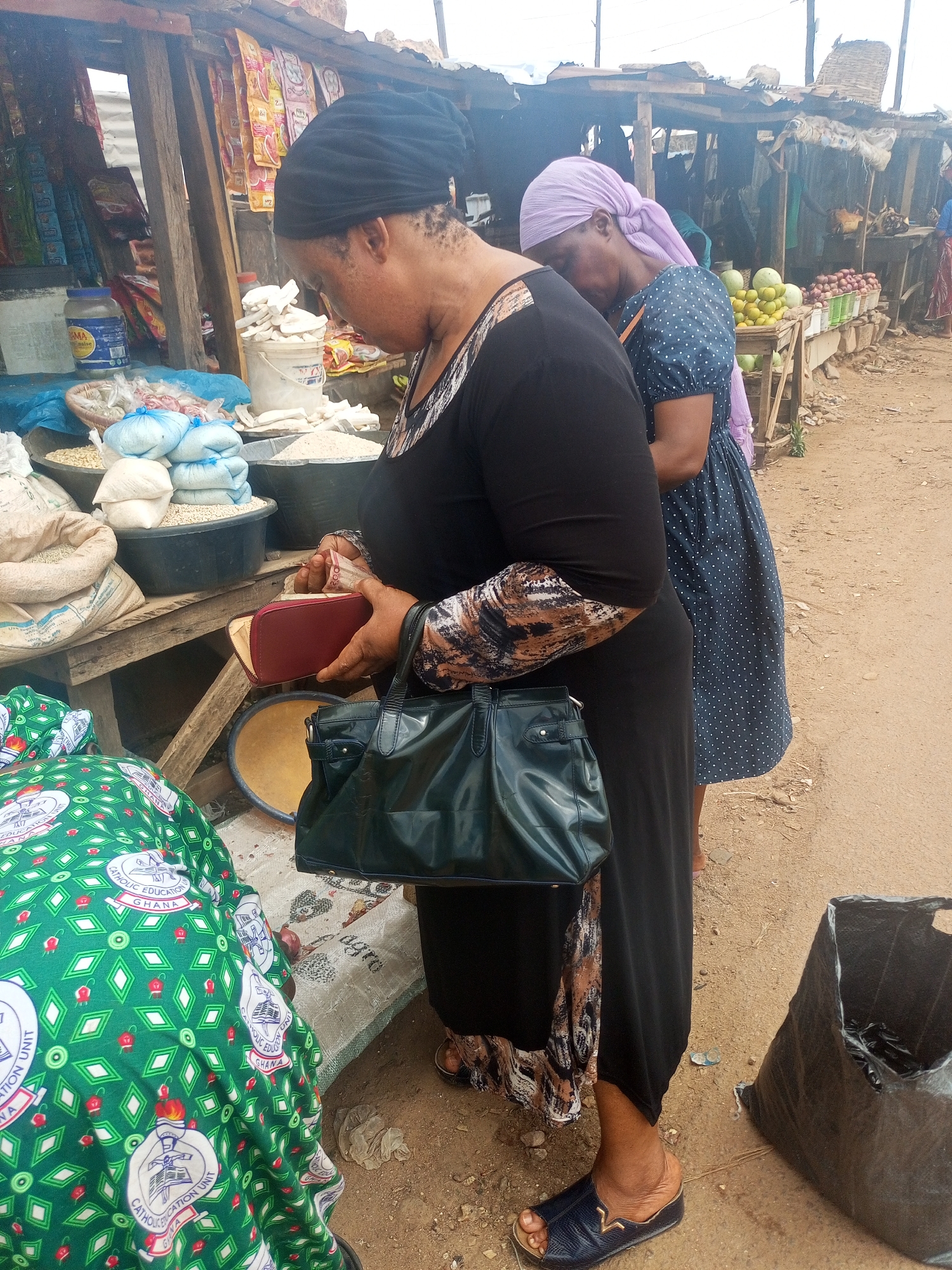 In an outdoor market in Sabon Tasha, Nigeria, a woman in a black dress and headscarf stands at a stall, examining the contents of a red wallet. She holds a dark green handbag. Bags and bins filled with grains and other products are on display. Behind her, another woman in a blue polka-dot dress and light purple headscarf stands by. The ground is dirt. To the left of the first woman is a green cloth with white and red text. Next to the first woman's feet, there is a black bag filled with items. The market stall is constructed from wood, and other stalls are visible in the background. Fruits and vegetables are on display at the stall in the background.