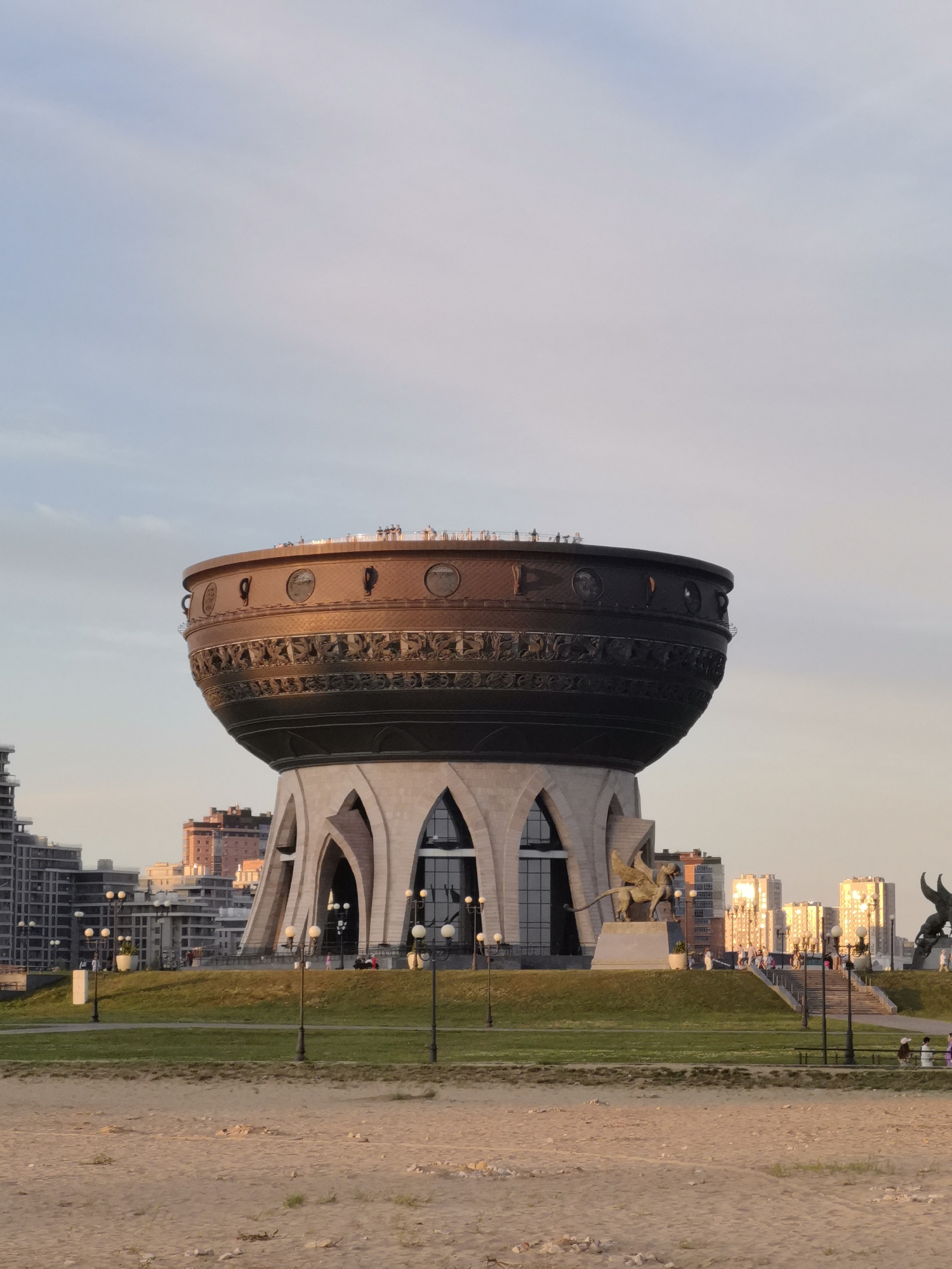 The image shows the Family Center "Kazan" in Kazan, Russia. The building is designed to resemble a giant cauldron, with a large, ornate copper bowl atop a base with arched windows. Several people are visible on the rooftop observation deck of the building, and a few people are walking on the grounds in front of it. In the background, there are several modern buildings, some of which are illuminated by the setting sun. The foreground of the image features a sandy area, likely a beach or riverbank, with some sparse vegetation. The sky is a pale blue with some wispy clouds.