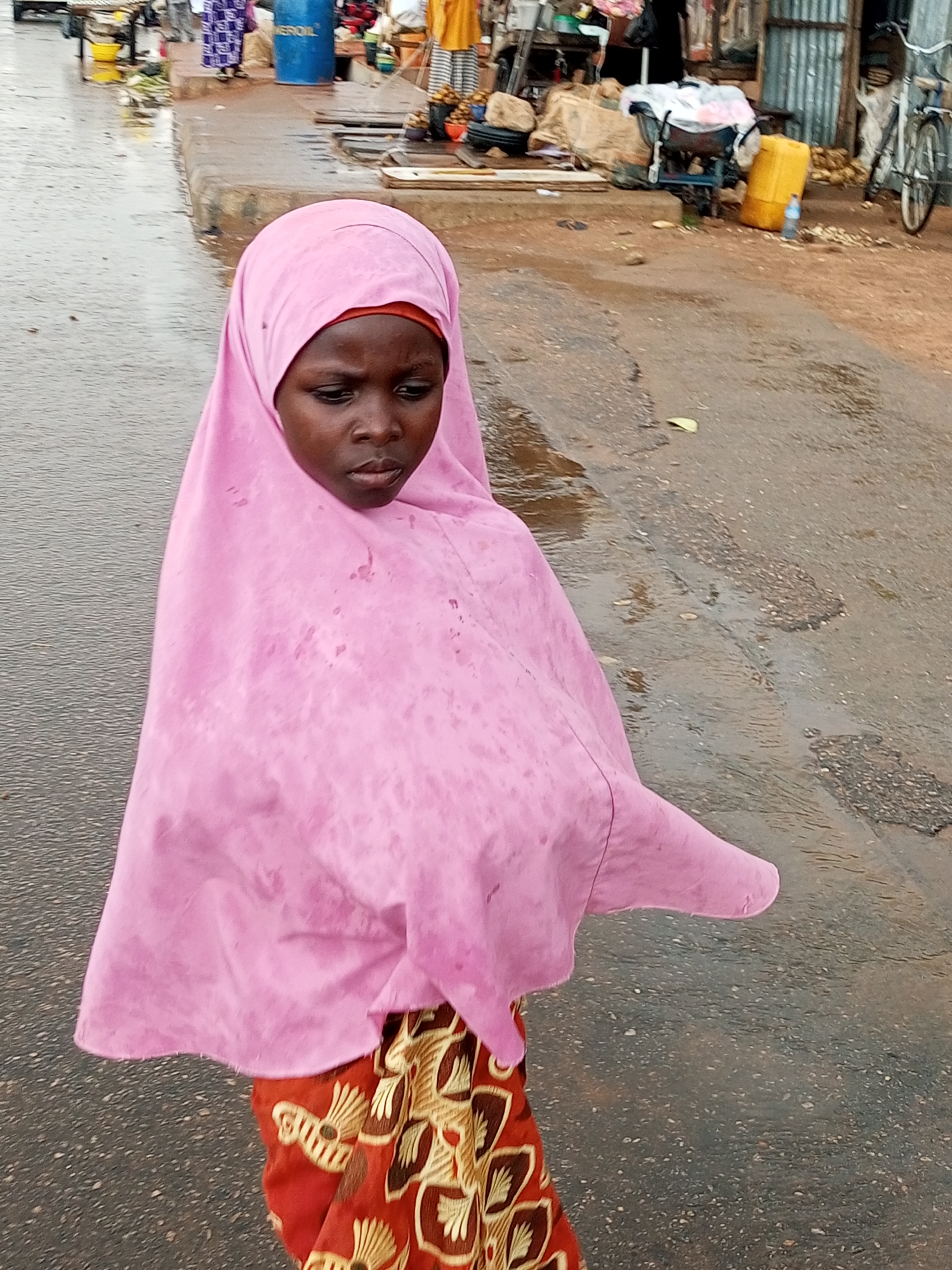 A young girl wearing a pink hijab and a patterned skirt stands on a wet, paved street in Tempure, Nigeria. The street is lined with market stalls and various objects, including barrels, crates, and a bicycle. The ground is wet and reflects the overcast sky, suggesting it may have recently rained or is currently drizzling. The girl is looking down and to her right, with a neutral expression. The scene appears to be a typical day in a bustling Nigerian marketplace.