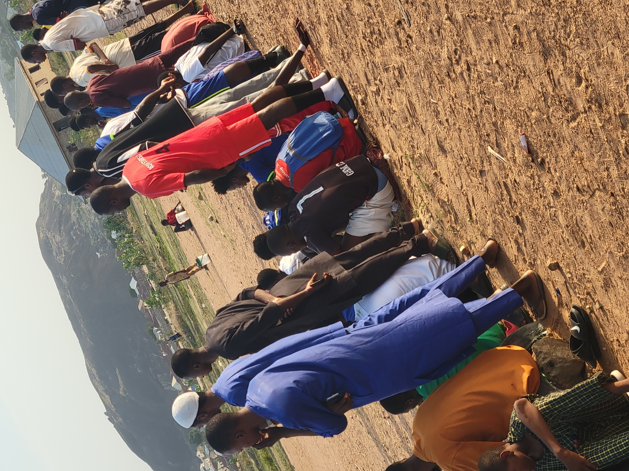 A group of approximately 15 men are lying prone on a sandy, open area in what appears to be Ibo, Nigeria. They are arranged in a line, with some individuals wearing traditional Muslim clothing such as a blue robe and white kufi cap. Others are in casual wear including t-shirts and shorts, with one wearing a red jersey with "ROYSTARS" printed on the back. They are lying face down, possibly engaged in prayer or some other form of communal activity. Behind them, a hilly landscape is visible, along with buildings and vegetation. The ground is mostly bare, sandy soil. The angle of the photo suggests it was taken from a slightly elevated position.