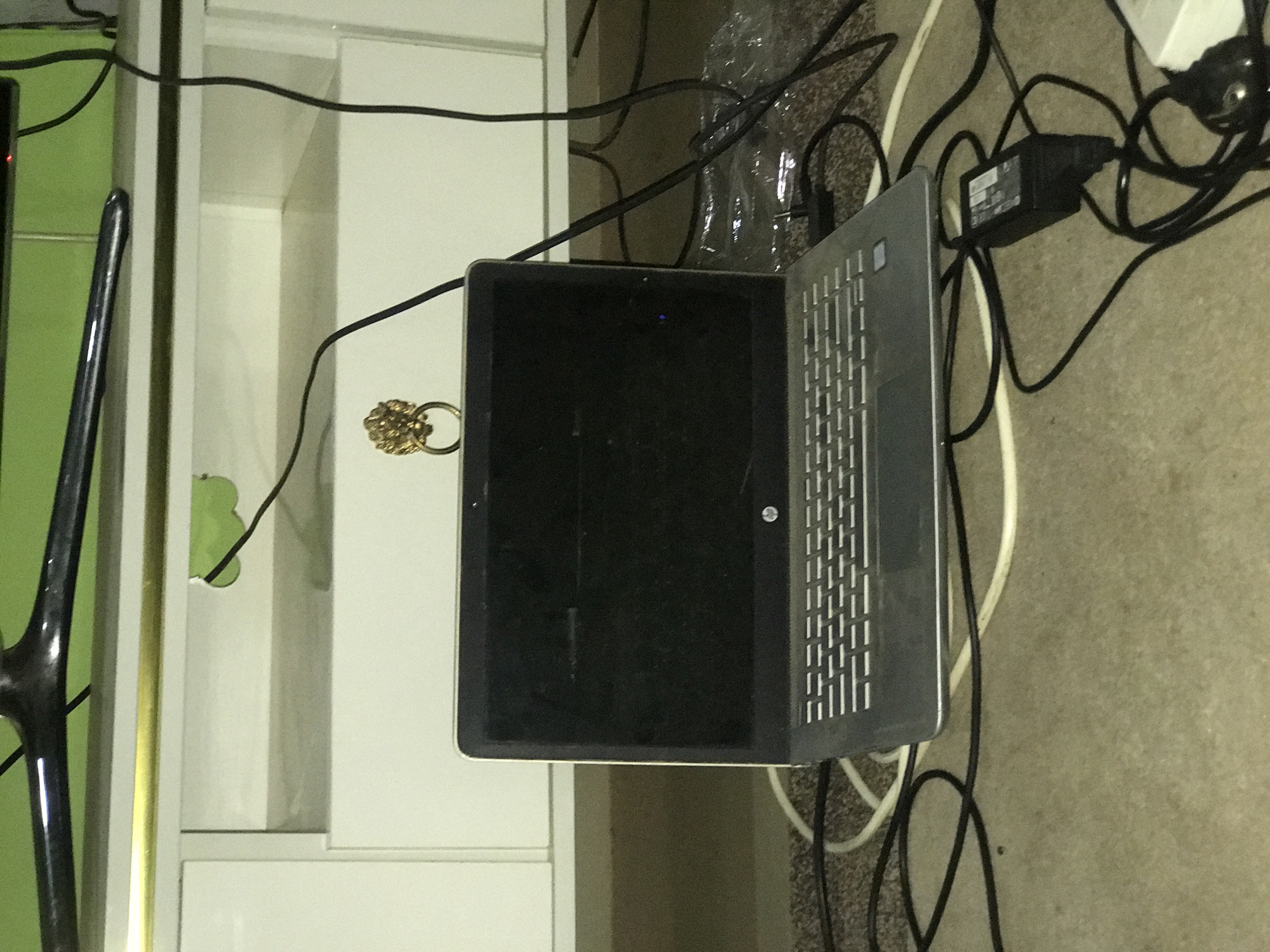 This image, taken in what is contextualized as Abuja, Nigeria, shows a cluttered indoor scene featuring electronics and furniture. The main subject is an open, silver-grey laptop resting on a light-colored, possibly carpeted, floor. Its screen is dark, but a small blue indicator light is visible near the top, suggesting the device is powered on or in standby.
Numerous black and white electrical cables are tangled across the floor and around the laptop. A black power adapter block, likely belonging to the laptop, is visible amidst the wires, along with a partially obscured white power strip or wall outlet. To the left of the laptop, there is a piece of white furniture, possibly a desk or cabinet, featuring shelves and a lighter top surface. Further to the left, a bright green surface or object is visible, along with a dark, glossy, curved metallic stand, potentially for a television or monitor. A small, ornate golden ring-like object hangs from a black wire near the laptop, appearing to be a decorative charm or keyring. A crumpled, clear plastic bag is also visible among the cables in the upper middle section.
No people are present, and no specific activity is taking place. The time of day, weather, and emotions are not discernible from the image. Text on the power adapter is too small to be legible.
