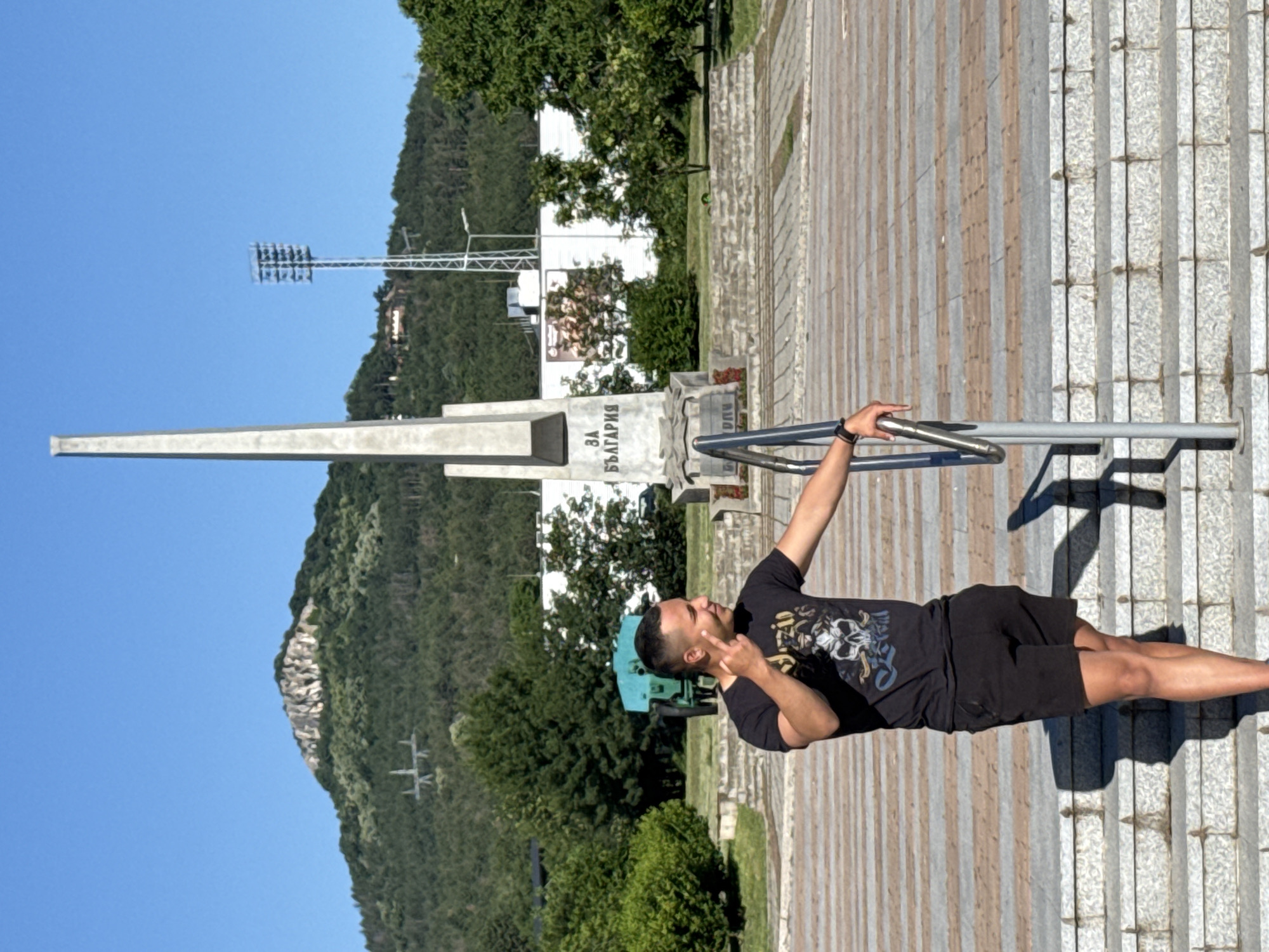 A young man with short hair, wearing a black t-shirt with a graphic print and black shorts, stands on a set of stairs, leaning on a metal railing. He is looking to his left and holding up two fingers in a peace sign. In the background, a tall, slender monument stands in front of a tree-covered hill. There is a stadium light on a tall pole to the right of the monument. A turquoise piece of machinery is visible behind the man. The ground is paved with patterned tiles, and the stairs are made of stone. The sky is clear and blue, and the sun is casting shadows.