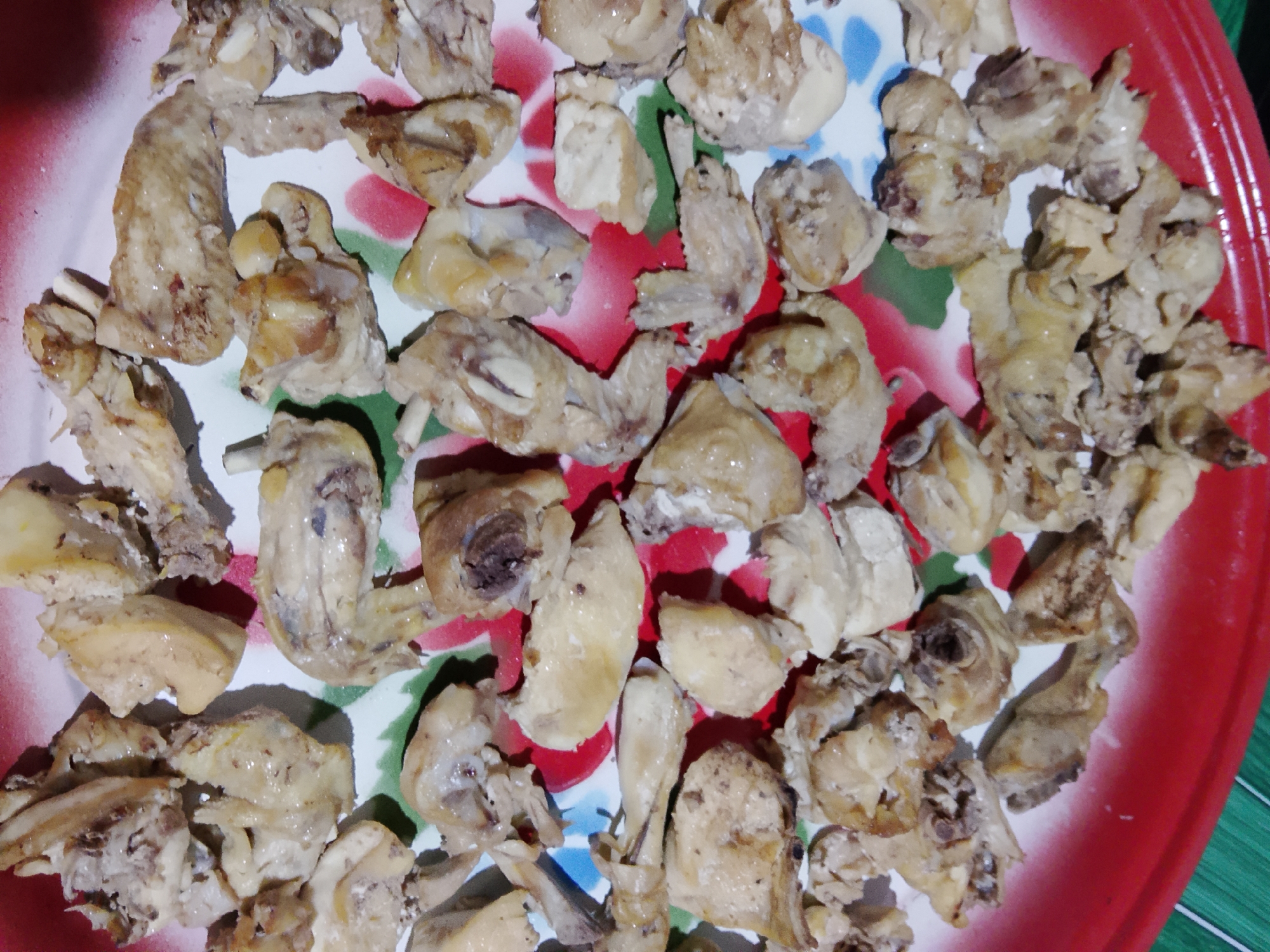 The image shows a close-up, overhead view of a large, round, red metal tray filled with numerous pieces of cooked chicken. The chicken pieces are varied in size and shape, appearing to be disjointed portions of a whole chicken, possibly including wings and small bone fragments. The chicken is light brown and appears slightly oily. The tray has a decorative pattern with red, white, green, and blue floral motifs, though the pattern is largely obscured by the chicken. The tray is placed on a surface with a green and white striped texture, likely a tablecloth or placemat. There are no people visible in the image. The scene is likely a food preparation or serving setting in Merbopuntong, Indonesia, where this dish is being presented.