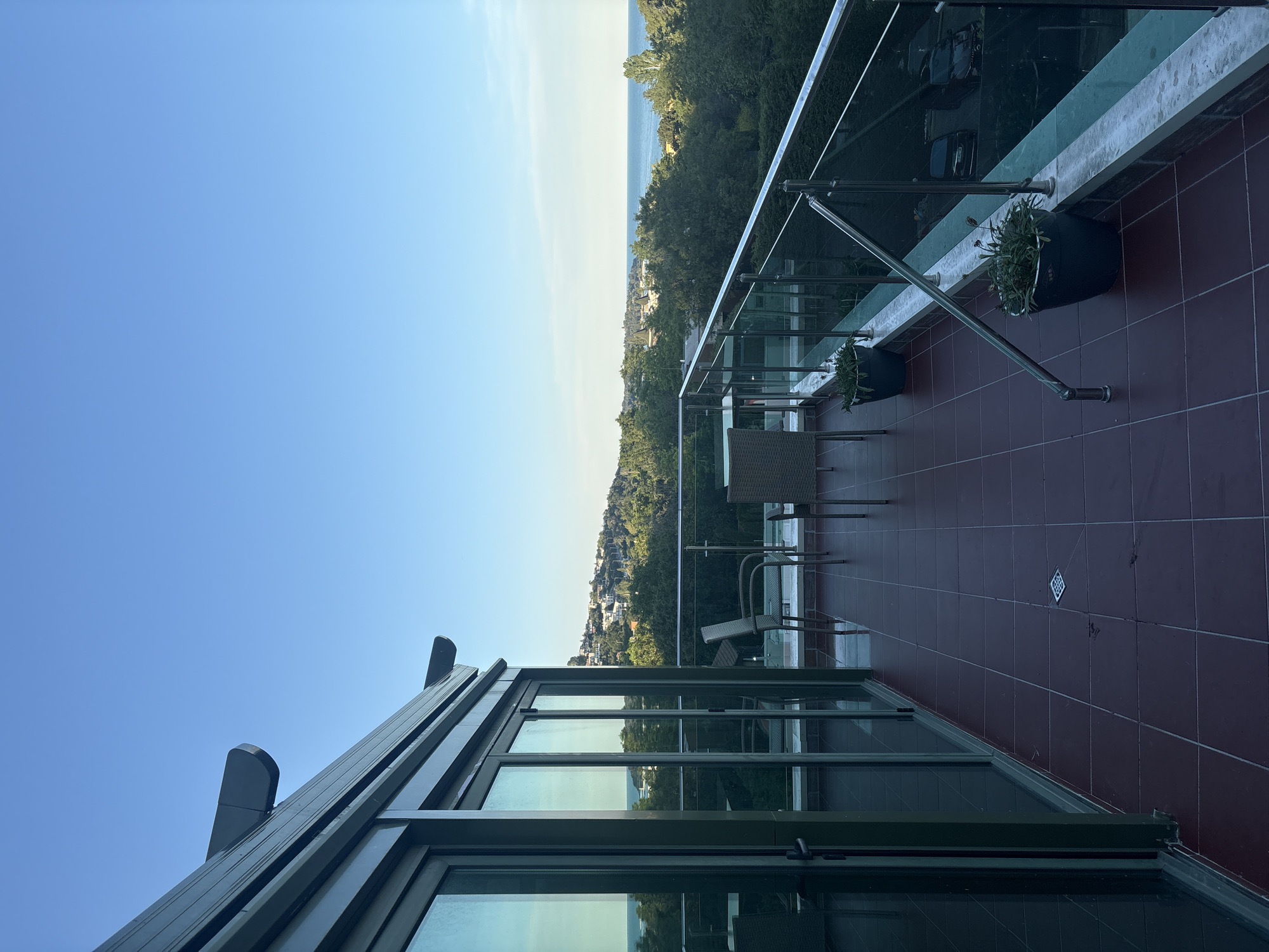 The image shows a balcony with a view of the sea and a town on a hill. The balcony floor is tiled in a reddish-brown color. There are two chairs and a table on the balcony, made of woven material. A glass railing with metal supports runs along the edge of the balcony. Two potted plants are placed on the balcony floor. Large glass doors lead from the balcony into an interior space. The sky is clear and blue, and the sun is shining. In the distance, the sea is visible, with a town nestled on a green hillside. Trees and other vegetation are abundant. The overall impression is one of a peaceful and scenic location.
