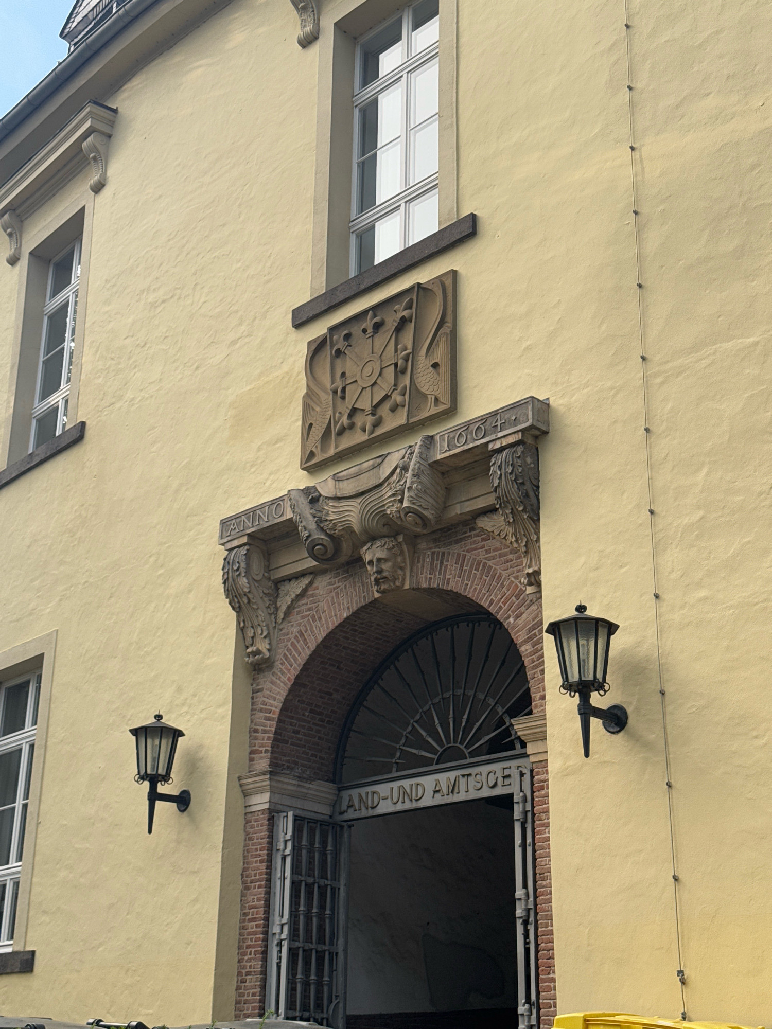 The image shows the entrance to the Land- und Amtsgericht (District and Municipal Court) in Cleves, Germany. The building has a pale yellow facade. Above the arched entrance, an inscription reads "ANNO 1604" which indicates the year of construction. The entrance itself is made of brick and is covered by a large metal gate. Two black, ornate wall-mounted lanterns are visible on the facade, one to the left and one to the right of the entrance. There are also two windows visible on the upper floor, one to the left and one to the right of the main architectural detail above the entrance. The overall impression is of an old, historic public building.