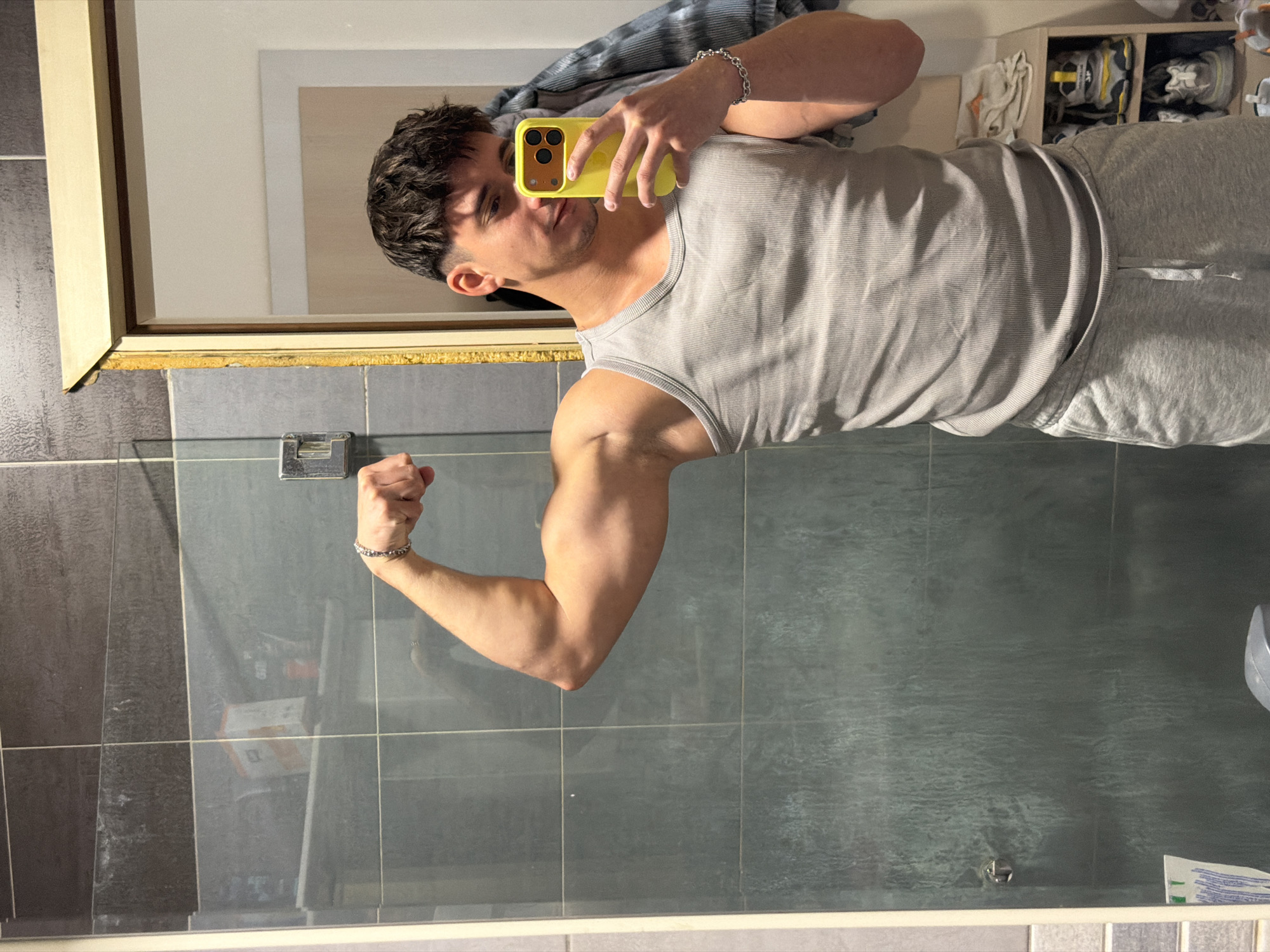 A young man is posing in front of a bathroom mirror, flexing his right arm to display his bicep. He is wearing a grey tank top and grey sweatpants. He's holding a yellow iPhone in his left hand, presumably taking the picture. The bathroom has grey and dark grey tiles on the wall. There is a mirror with a gold frame above the sink and on the top right of the image there is a shelf. The location cannot be determined from the image.