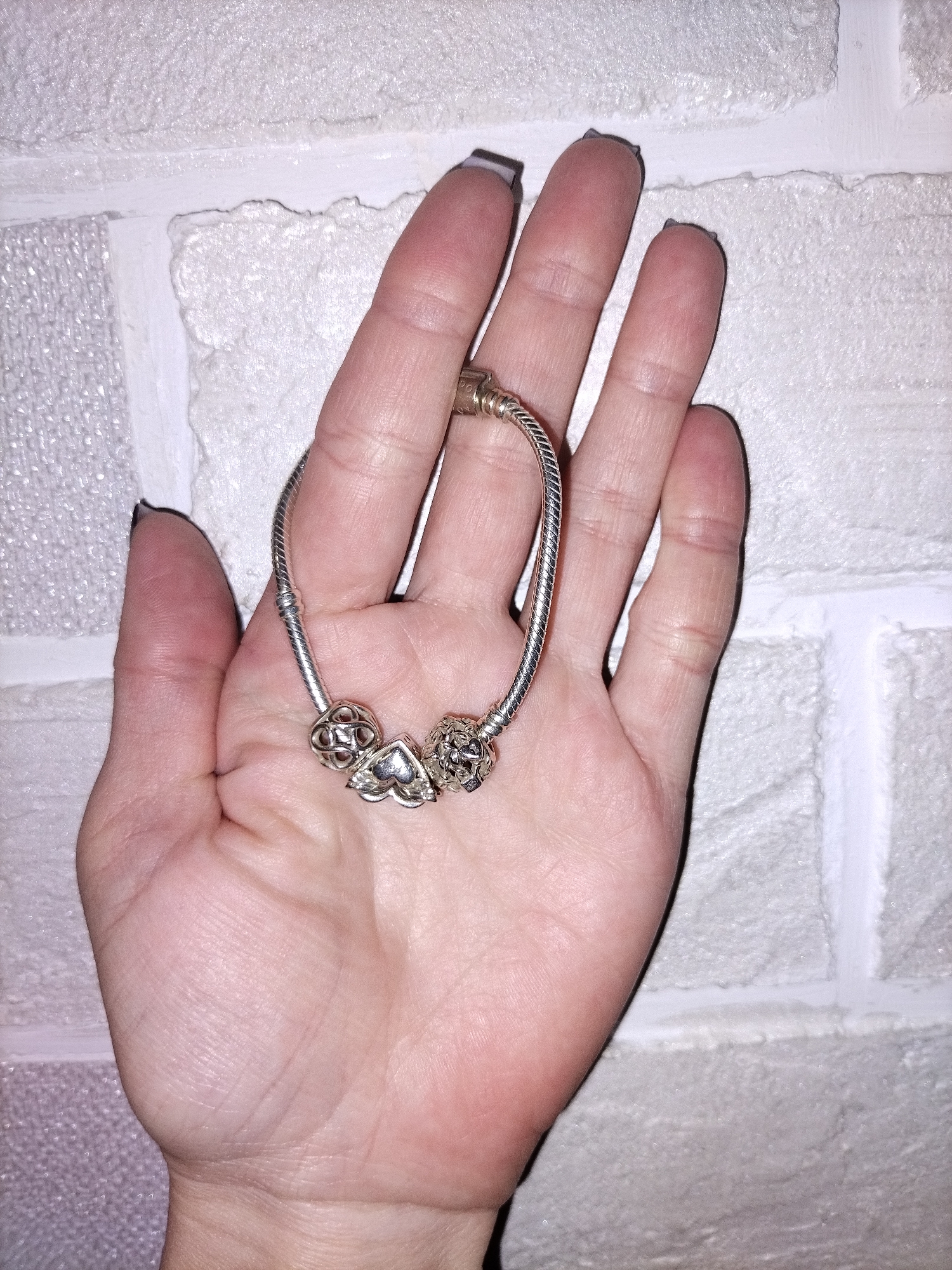 A human hand, palm facing upwards, holds a silver charm bracelet against a textured, light-colored wall. The hand displays visible palm lines and creases, and fingernails are painted with dark polish. A gold-colored ring is partially visible on one finger.
The bracelet features a braided silver chain adorned with three distinct charms. One charm is a stylized heart shape, another is a spherical charm with intricate filigree or openwork, and the third is a spherical charm with a geometric or knot-like pattern. The silver material of the bracelet and charms reflects the ambient light.
The background is a wall with a rough, uneven surface, resembling whitewashed bricks or stone, with visible grout lines. Portions of the wall, particularly on the left side, exhibit a subtle sparkly or granular texture, suggesting a specific type of paint or wallpaper. The lighting appears to be indoor, casting soft shadows on the hand and bracelet. There is no visible text in the image.