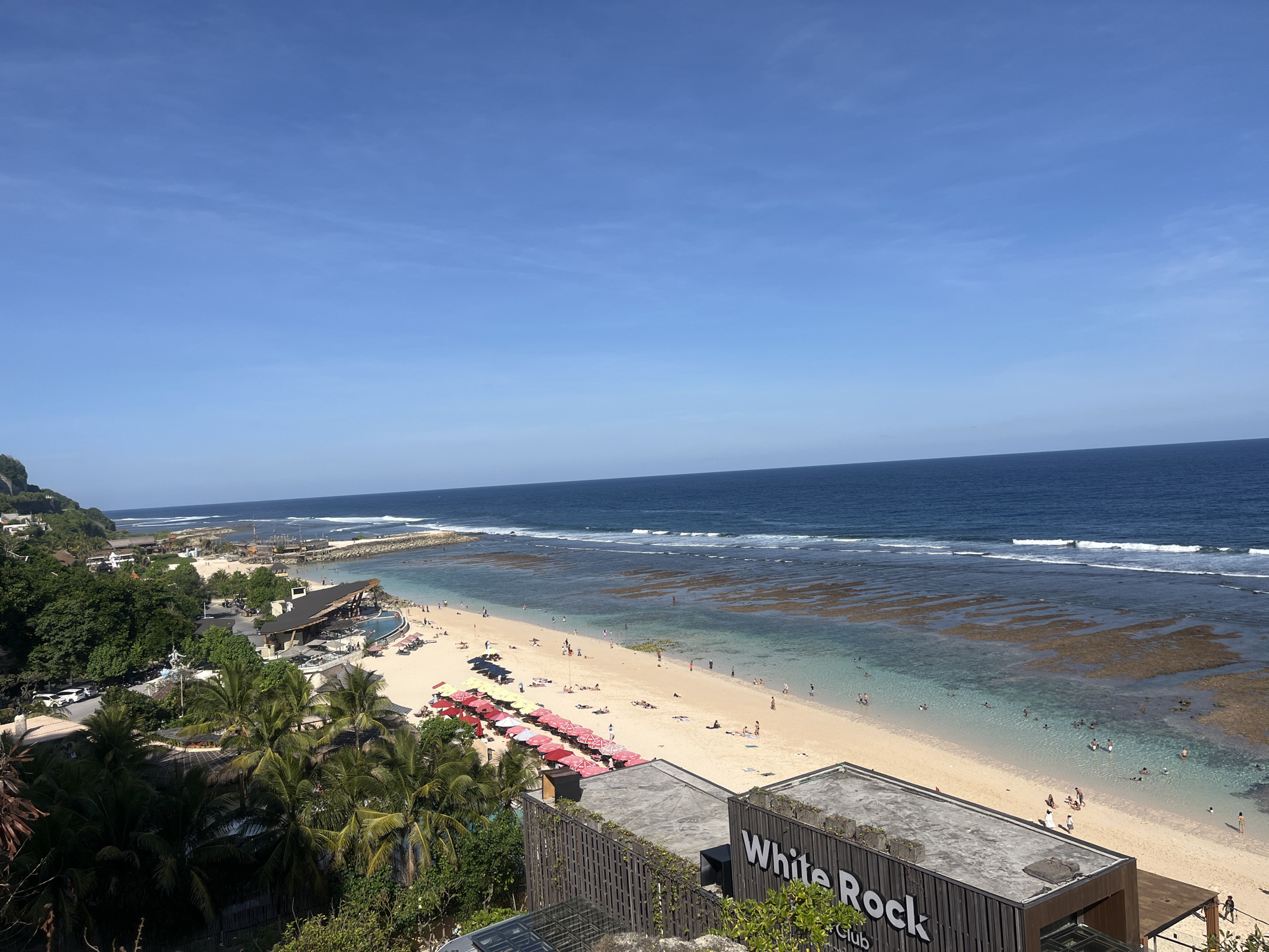 The image shows a sunny beach scene in Kuta Selatan, Indonesia. A sandy beach stretches along the coastline, dotted with people sunbathing, swimming, and walking. The ocean is clear and shallow near the shore, with visible coral or rock formations beneath the surface. Waves are breaking further out at sea. A row of red and pink umbrellas line the beach. In the foreground is a building with "White Rock Club" displayed on its side. Palm trees are visible in the lower left corner of the image. The sky is clear and blue.
