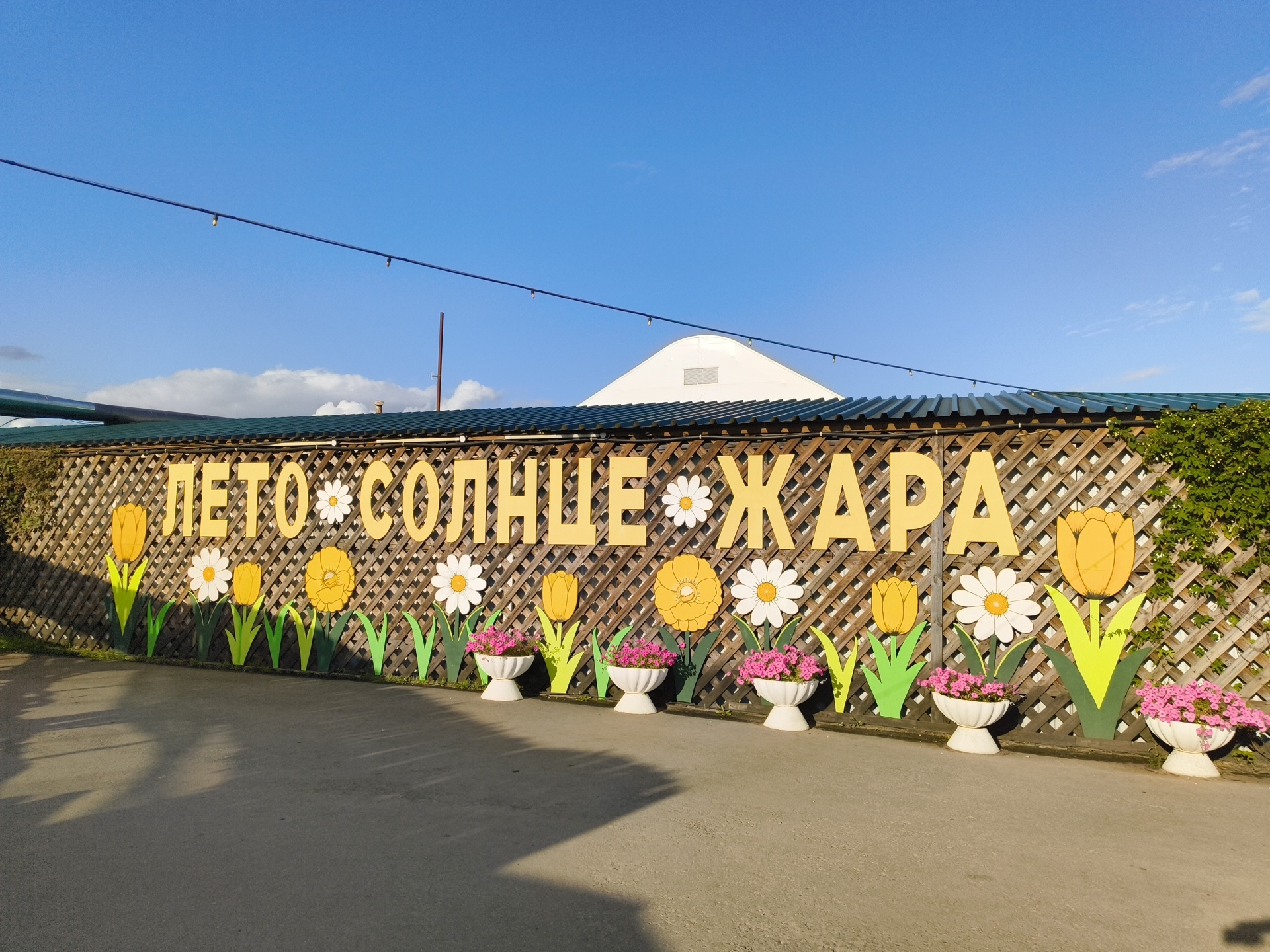 The image shows a wooden lattice fence decorated with large yellow letters spelling "ЛЕТО СОЛНЦЕ ЖАРА" (Summer Sun Heat) in Russian. Below the lettering, there are cutouts of colorful flowers, including yellow tulips and white daisies. White planters filled with pink flowers are placed along the base of the fence. In the background, a white building with a curved roof is visible, along with a clear blue sky. The scene appears to be outdoors, with a paved ground in the foreground. The image is likely taken in a public space in the Малахавецкі сельскі Савет, Belarus, possibly a park or a recreational area. No people are visible in the image.