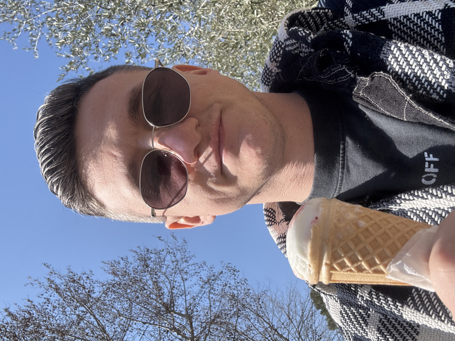 A close-up selfie captures an adult male outdoors in Peschiera del Garda, Italy, under a clear blue sky. The subject, with short, dark hair slicked back and visible graying at the temples, wears dark, hexagonal-framed sunglasses. He exhibits a neutral expression with a slight smile. His attire includes a dark t-shirt with the partial text "OFF" visible, layered under a black and white patterned, woven jacket. He holds a single scoop of white ice cream with reddish streaks in a standard waffle cone, partially wrapped in white paper, in his left hand, positioned at the lower center of the frame. The background features the top branches of a bare deciduous tree to the left and an olive tree with silvery-green leaves to the right, indicating a sunny environment.