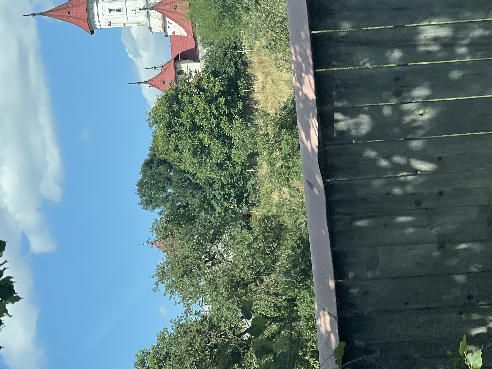 A wide shot captures a castle-like structure with red-tiled roofs and white walls, partially obscured by lush green trees and foliage. The castle has several pointed towers and decorative architectural elements. In the foreground, a dark wooden fence with visible vertical planks runs horizontally across the lower portion of the frame, casting dappled shadows from overhead leaves onto its surface. The background is a clear blue sky with scattered white clouds. The scene suggests a sunny day in a rural or park-like setting, possibly near Capu Codrului, Romania, given the castle's architectural style. There are no people or animals visible in the image.