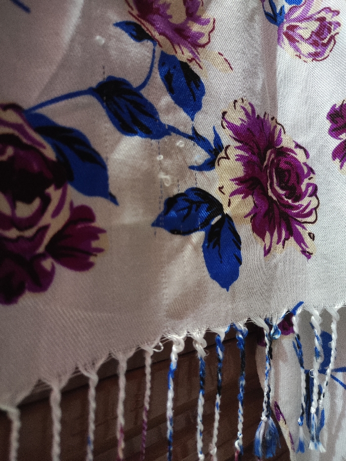 The image presents a detailed, close-up view of a light-colored textile with a repeating floral pattern, located in Astanajapura, Indonesia. The fabric displays large, stylized roses with petals in shades of purple and magenta, accented by cream or yellow, and accompanied by dark blue leaves with black outlines. The material exhibits a visible woven texture, with small, light-colored fiber nubs present on its surface. The bottom edge of the textile is finished with a fringe, consisting of multiple twisted strands that are predominantly white, with some incorporating blue and purple threads consistent with the fabric's pattern. The immediate background behind the fringe appears to be a dark, textured brown surface, consistent with wood, suggesting an indoor environment. No individuals, overt actions, or interactions are depicted in the scene.
