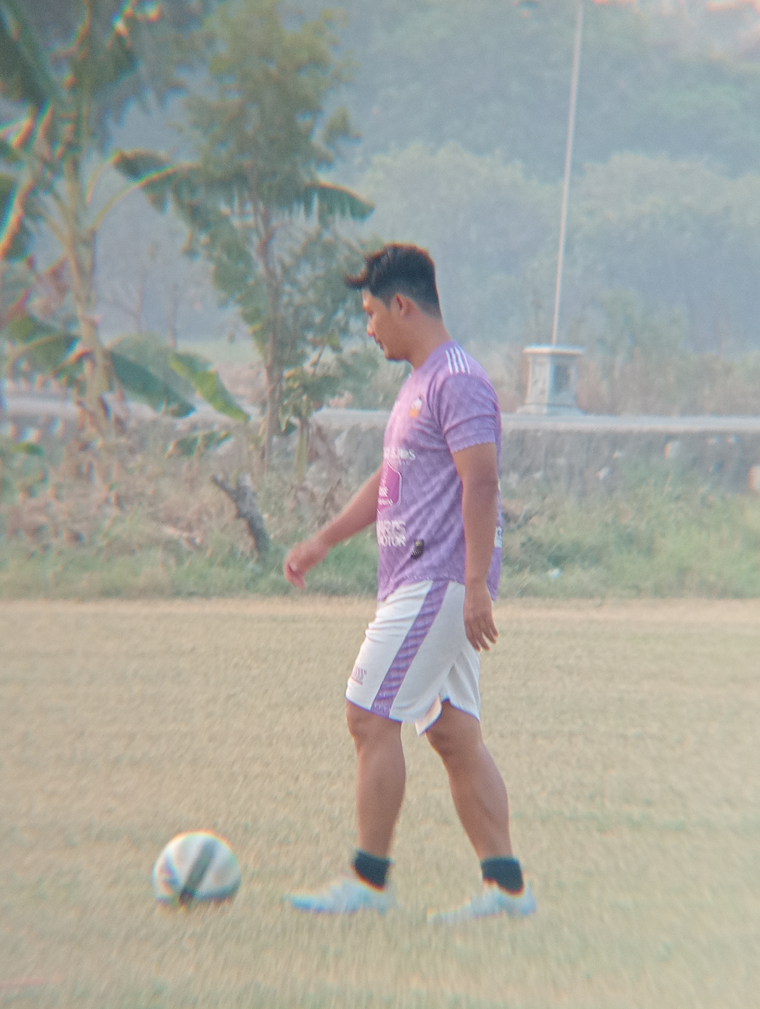 A man is walking on a grassy field, with a soccer ball at his feet. He is wearing a purple and white soccer jersey and shorts. The field appears to be in a rural area, with trees and buildings visible in the background. The sky is overcast, and the lighting is dim. The image was taken in Kenep, Indonesia.