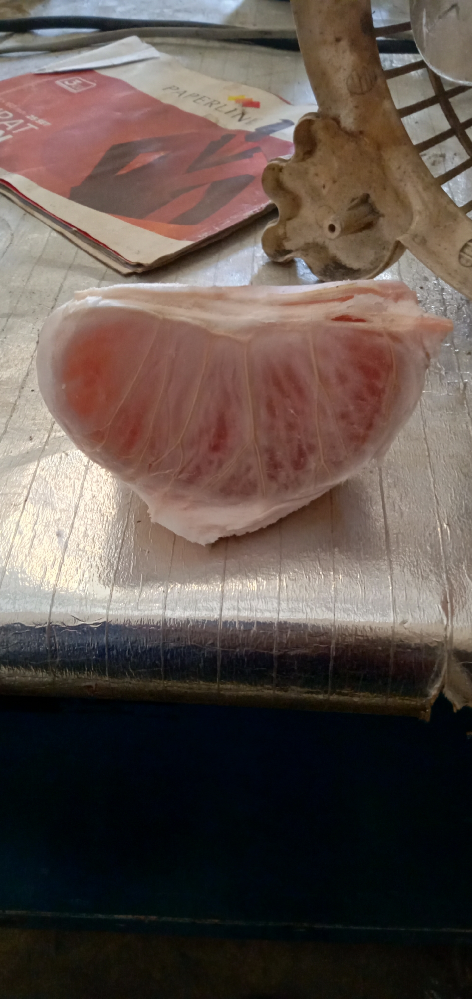 The image shows a segment of a peeled pink grapefruit on a corrugated silver surface, possibly a table or counter. In the background, there's a magazine with the word "PAPERLINE" visible on the cover, featuring a graphic design in red. A partially visible, possibly metallic object with a textured, star-shaped knob is also present in the background. The location is Sumput, Indonesia.