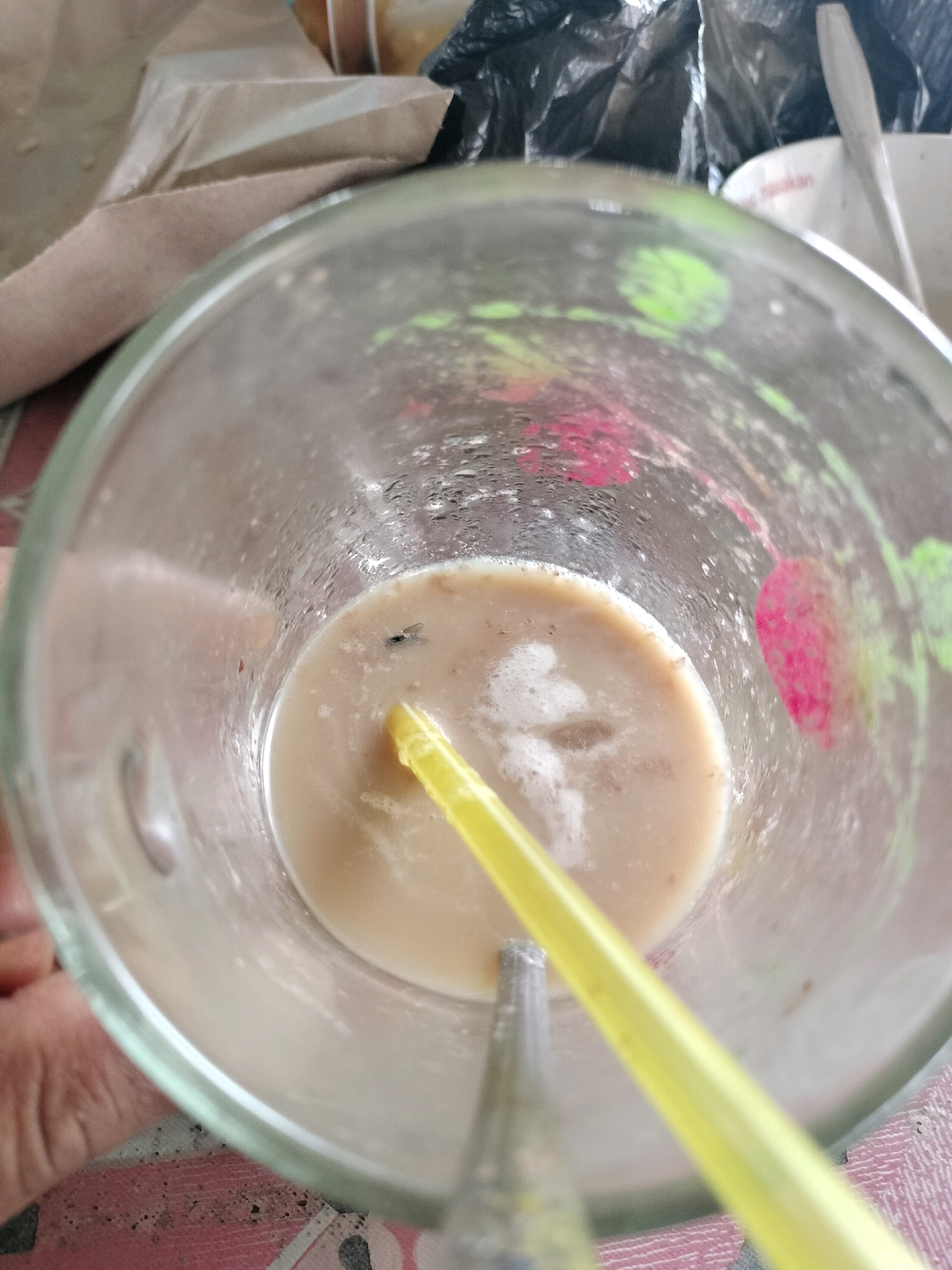 A fly is resting on the surface of a milky beverage in a clear glass mug. A yellow straw extends from the bottom of the mug and slightly into the liquid. The mug is sitting on a patterned surface, possibly a table. In the background, there are various items, including brown paper packaging and black plastic bags. The scene appears to be in an outdoor setting, possibly a street-side stall or cafe in Maburai, Indonesia, given the context provided. There are no people visible in the image.