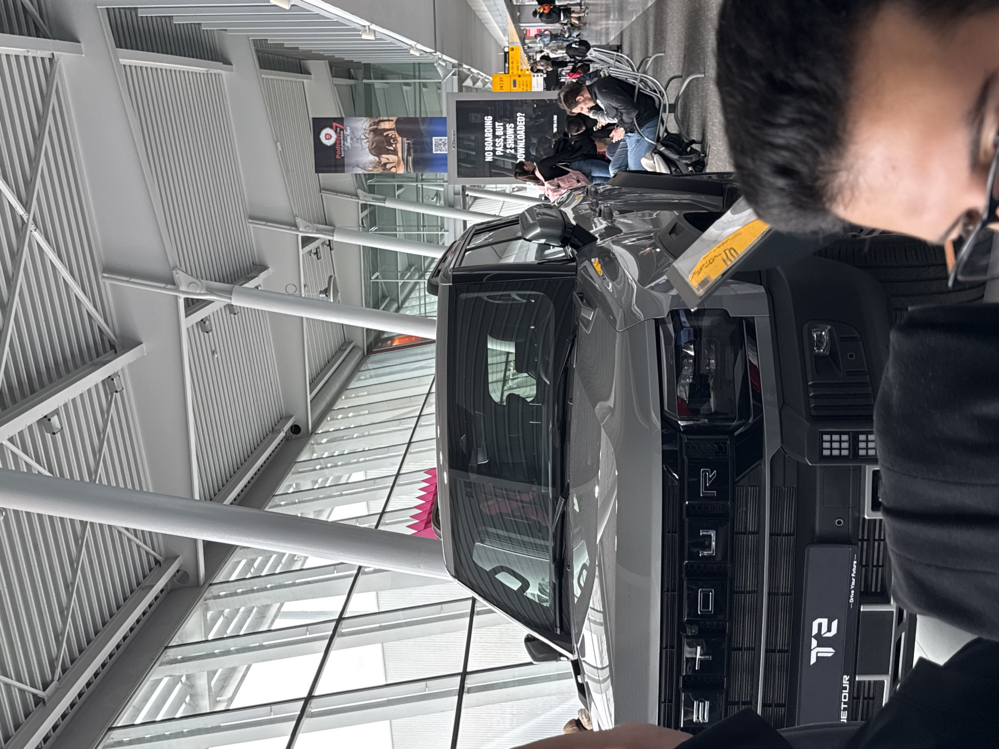 The image depicts an indoor scene within a modern airport terminal in Warsaw, Poland. A large, grey SUV or truck is prominently displayed in the center, acting as a focal point. The vehicle features the branding "JETOUR" and "T2" on its grille, along with the slogan "-Drive Your Future->."
In the immediate foreground, the back of a person's head and shoulder are visible, out of focus, suggesting they are observing or photographing the display vehicle. This individual wears glasses. Behind the SUV, several other people are visible, seated on airport benches or standing, indicative of passengers waiting in a concourse area. Some appear to be looking at their phones or interacting with each other.
The setting is characterized by contemporary airport architecture, featuring high ceilings with exposed metallic support beams and corrugated panels. Large windows allow natural light to illuminate the space, suggesting it is daytime.
Several advertisements are visible in the background. One large black banner asks, "NO BOARDING PASS, BUT 2 SHOWS DOWNLOADED?". Another banner, featuring wildlife (likely deer or similar animals), displays the text "Podróże na kontynentów" (Polish for "Journeys to continents") and the "JCDecaux" logo, along with a QR code. Yellow signs showing the numbers "28 27" are also present, likely denoting gate numbers. The general ambiance is bright and busy, typical of an airport environment.