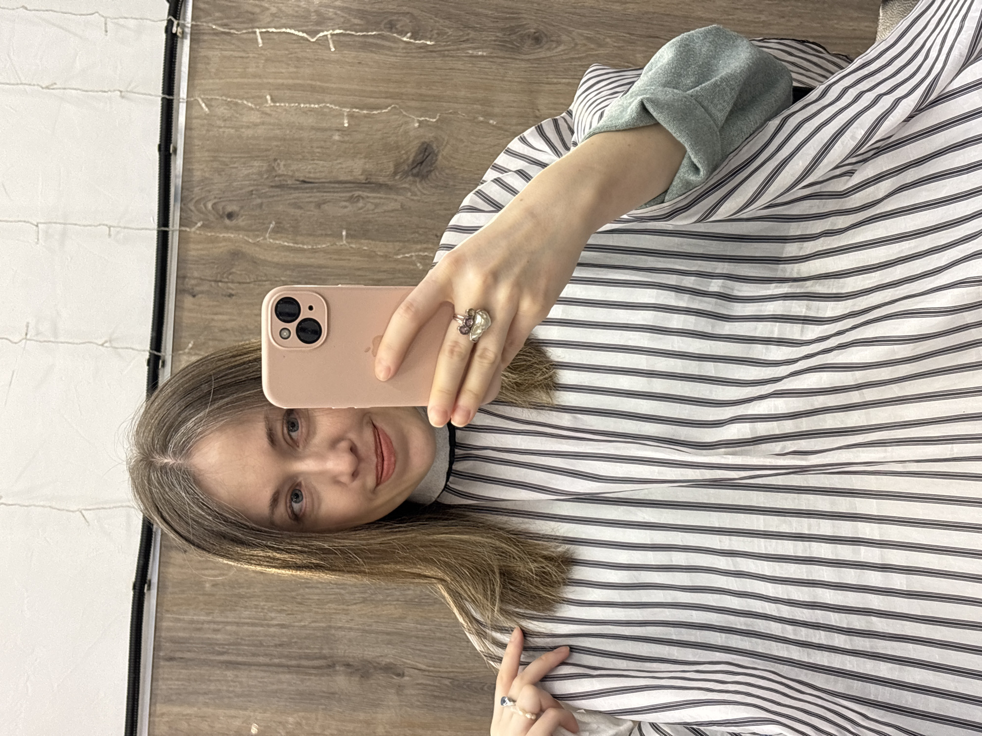 A woman is taking a selfie in what appears to be an indoor setting. She is holding a pink smartphone, likely an iPhone, in her right hand. She is wearing a striped shirt, possibly a smock, over a gray sweatshirt or top. Her hair is blonde with visible gray roots. She has light skin and blue eyes. There is a ring on her finger. The background is a combination of a wood-like surface, which is possibly the floor, and a white surface, maybe a backdrop or wall. The lighting suggests an indoor environment, possibly a salon or home setting. No specific features indicate the location is Saint Petersburg, Russia.