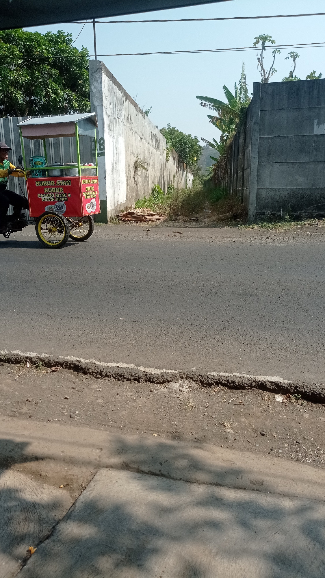 A side view of a person on a motorcycle pushing a food cart is captured on a sunny day in Bedali, Indonesia. The food cart is red and yellow and displays signage in Indonesian advertising "Bubur Ayam" (Chicken Porridge) and "Bubur Kacang Hijau & Ketan Hitam" (Mung Bean Porridge & Black Sticky Rice). The motorcycle and cart are on the right side of the road, moving away from the viewer. Behind the cart, a tall, rough concrete wall runs parallel to the road, with vegetation growing on either side. Trees and foliage are visible in the background, and the sky is clear and blue. The road itself is asphalt with visible cracks and wear along the edges. The foreground shows a concrete path with shadows cast by overhead objects, indicating a bright, outdoor environment.