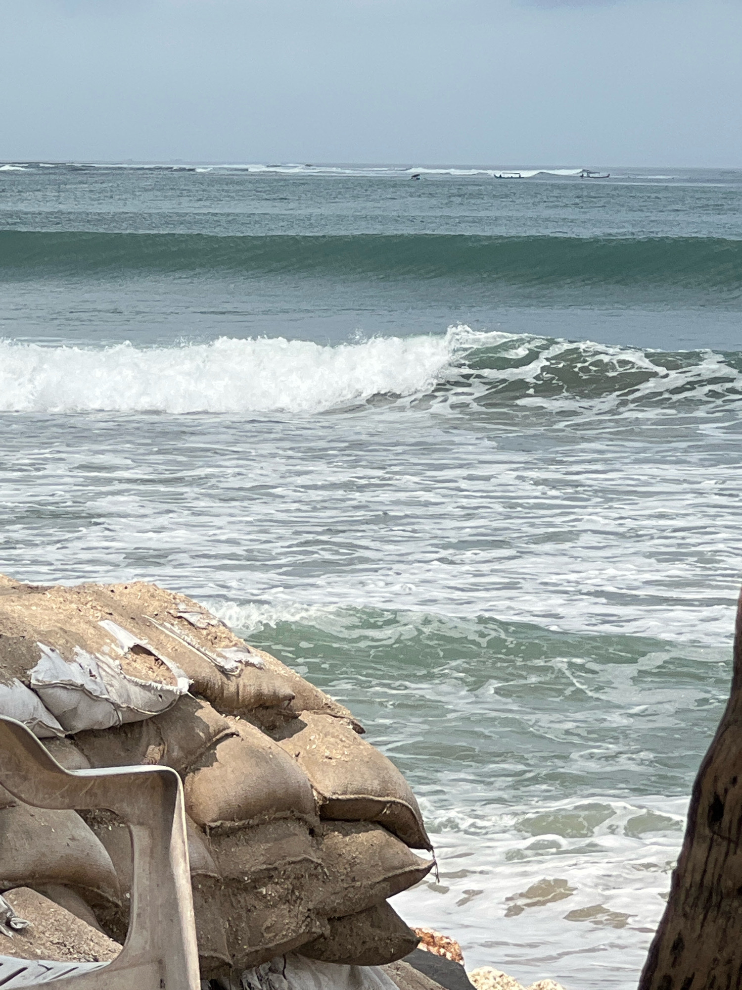 The image depicts a serene coastal scene with a prominent focus on sandbags and a partially visible plastic chair in the foreground. The sandbags, stacked in a weathered state, suggest a defensive barrier against the encroaching sea. The ocean stretches out in the background, with gentle waves rolling towards the shore. In the middle ground, several small boats are visible on the horizon, hinting at local fishing activities or perhaps recreational use. The overall environment is that of a beach with a muted color palette, under an overcast sky. The presence of sandbags and the visible waves suggest a location prone to coastal erosion or storm surges, which is common in areas like Kuta, Bali, Indonesia, known for its surf beaches and susceptibility to the elements.