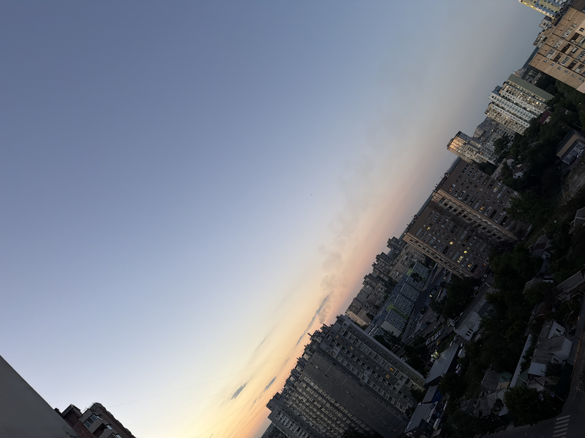The image is a high-angle, wide shot of a dense urban residential area in Kyiv, Ukraine, taken at sunset. The sky displays a gradient of colors, from pale yellow and pink near the horizon to a soft blue overhead, indicating the late afternoon or early evening. The buildings are a mixture of Soviet-era apartment blocks and some newer structures, all characterized by their height and numerous windows. Many of the buildings are tall, some appearing to be 15-20 stories high, and are tightly packed together.
In the foreground on the left, the corner of a building is visible, extending vertically upwards out of frame. This building appears to be older, with visible brickwork and windows. The rest of the image is dominated by the city skyline, with buildings stretching into the distance. Some buildings reflect the warm sunset light.
The urban environment is filled with the typical elements of a residential district: streets, parks or green spaces dotted with trees, and visible power lines crisscrossing between buildings. There are also parked cars visible on some streets and in courtyards. The perspective is tilted, giving a sense of looking down and across the city from a high vantage point. The scene is devoid of visible people or any discernible activity. The overall impression is one of a typical residential neighborhood in a large Eastern European city during twilight.