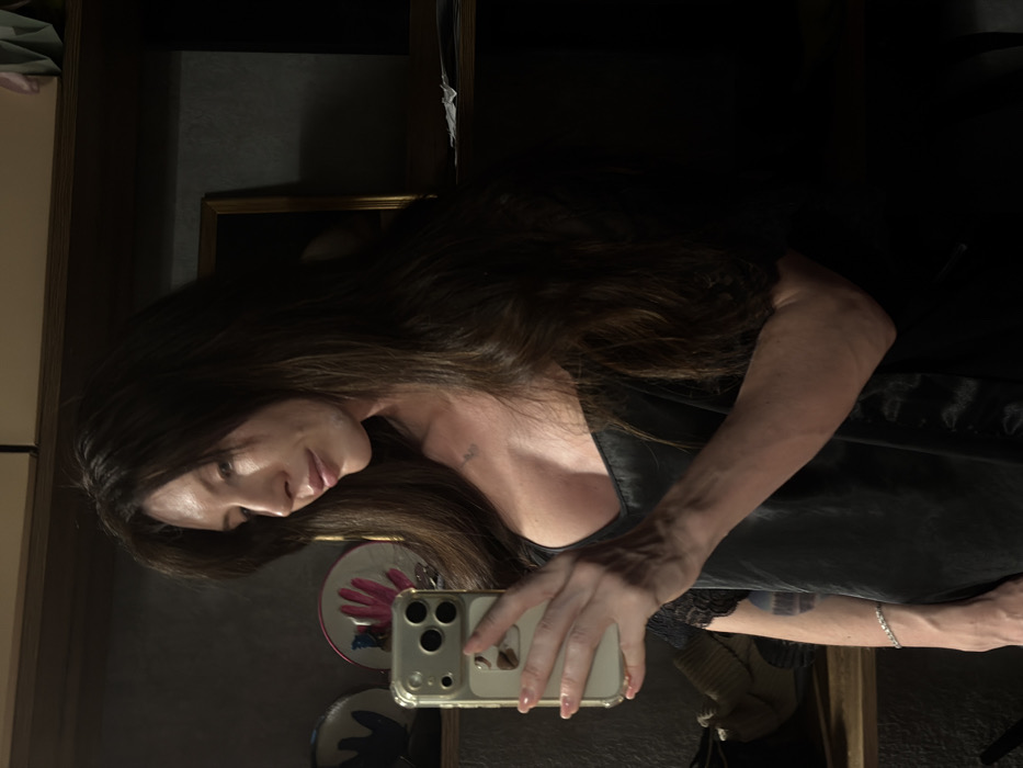 A woman with long brown hair is captured in a mirrored selfie. She's holding a phone, its reflection revealing part of the room, including a patterned plate. She wears a dark, low-cut top. The background is dim, featuring a cabinet and a hint of decorative elements.
