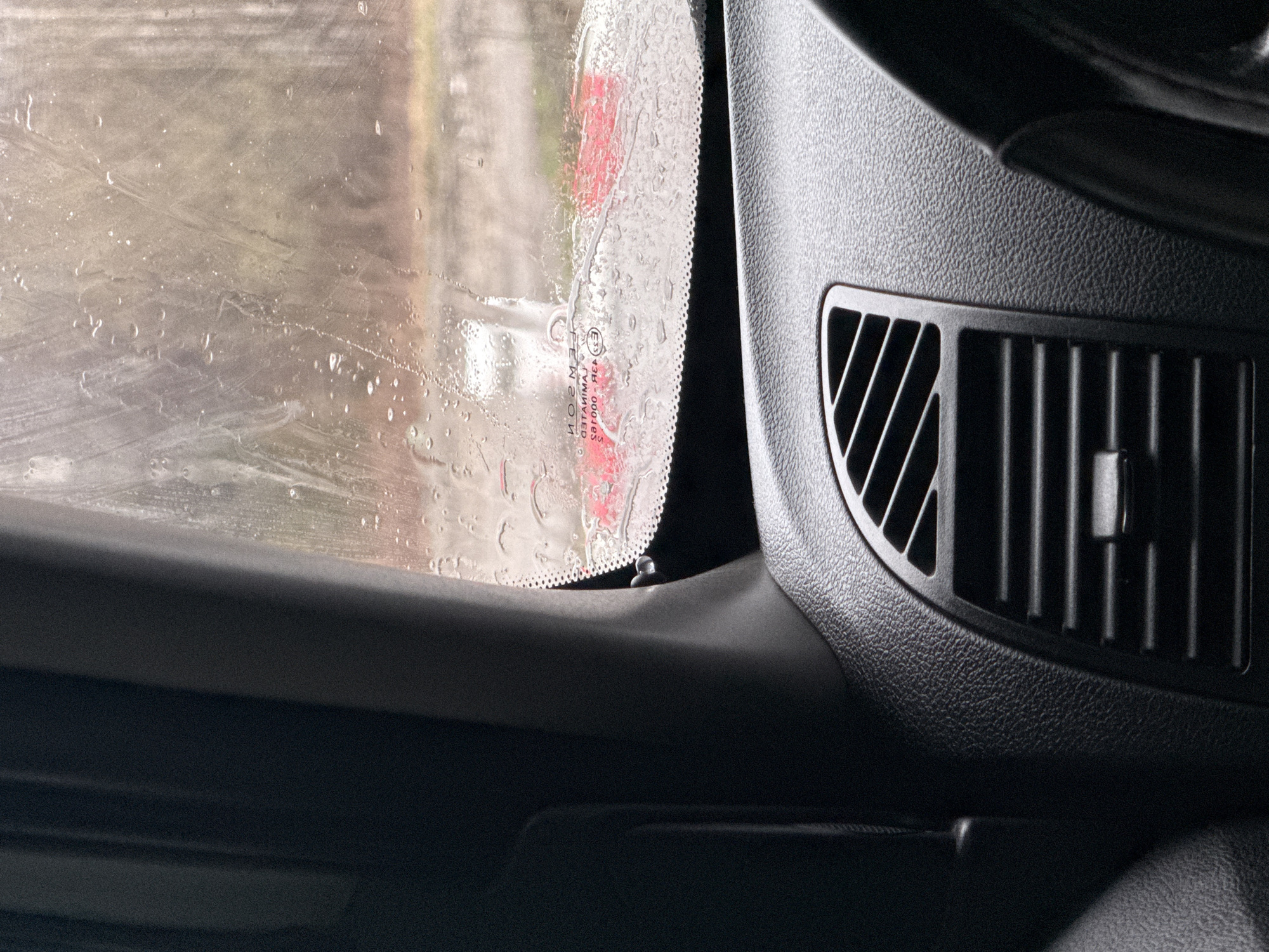 This close-up image, taken from inside a vehicle, captures a wet car window and a section of the dashboard. The left side of the frame is dominated by the car's window, which is heavily streaked and dotted with water droplets, suggesting either rainfall or a car wash. Through the blurred, wet glass, a muted, indistinct outdoor environment is visible, with a faint red hue reflecting or appearing in the distance.
Along the edge of the window, a black frit band is discernible, and mirrored text indicating the glass manufacturer and safety standards is visible. The text, viewed from inside, reads "LAMINATED," "SAINT-GOBAIN SEKURIT," "E2," and "43R-000105." This implies the window is European-standard laminated safety glass.
The right side of the image showcases a dark, textured plastic surface of the car's dashboard. A vertical air conditioning or heating vent with multiple slats is prominently featured in the lower right quadrant. The overall lighting suggests an overcast day or an interior shaded from direct sunlight, with the exterior appearing brighter but diffused. No people are visible, and the scene implies the vehicle is either stationary during precipitation or undergoing a wash in Klenovskoe, Russia. The focus on the water droplets and the interior details creates an intimate perspective from within the car.