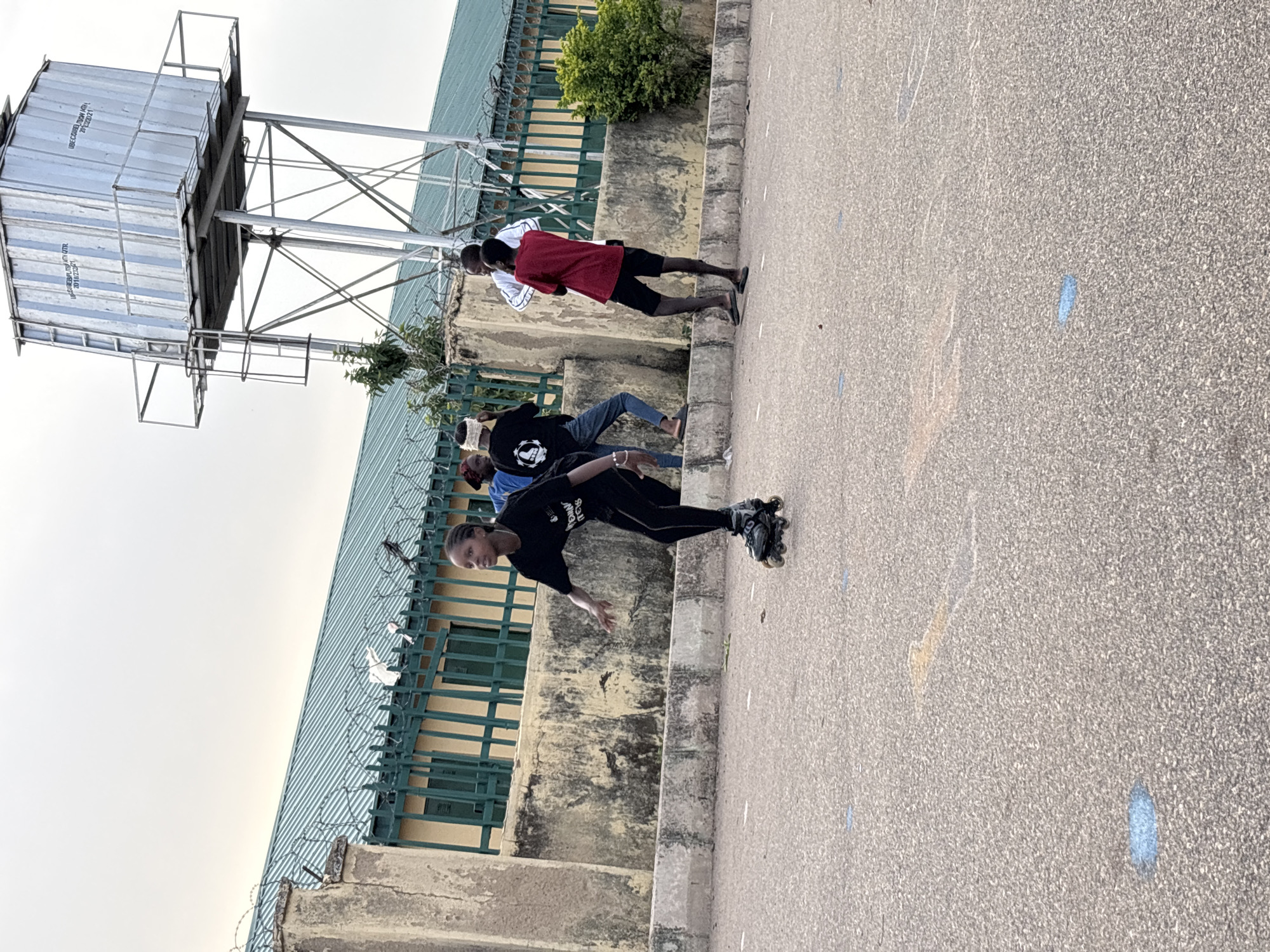 The image, rotated 90 degrees clockwise, captures an outdoor scene featuring several individuals on a paved area, likely in Jos, Nigeria.
In the foreground, a young person, possibly female, dressed in black athletic wear and rollerblades, is actively skating, leaning to one side. The asphalt ground beneath them features scattered blue and faded white markings.
In the midground, several other people are present. One individual wearing a red shirt and black shorts stands near a low concrete barrier, being observed by another person in a white shirt. Further back, another person in a black t-shirt and jeans is partially visible, appearing to be resting or watching the activity.
The background features a long, low building with a green corrugated metal roof and yellow-painted walls, protected by a metal fence topped with barbed wire. Green shrubbery grows along the base of the building.
Prominently featured in the upper left (when correctly oriented) is a large, elevated metal water tank, resembling a repurposed shipping container, supported by an intricate metal truss structure against a bright, possibly overcast, sky. Text visible on the tank includes "IBC CARGO TRAIN AND..." and "2018/2019", along with other partial lettering.
The overall activity depicted is roller skating or rollerblading, suggesting a casual recreational gathering or practice session. The lighting indicates daytime, possibly late afternoon, with soft, diffused light.