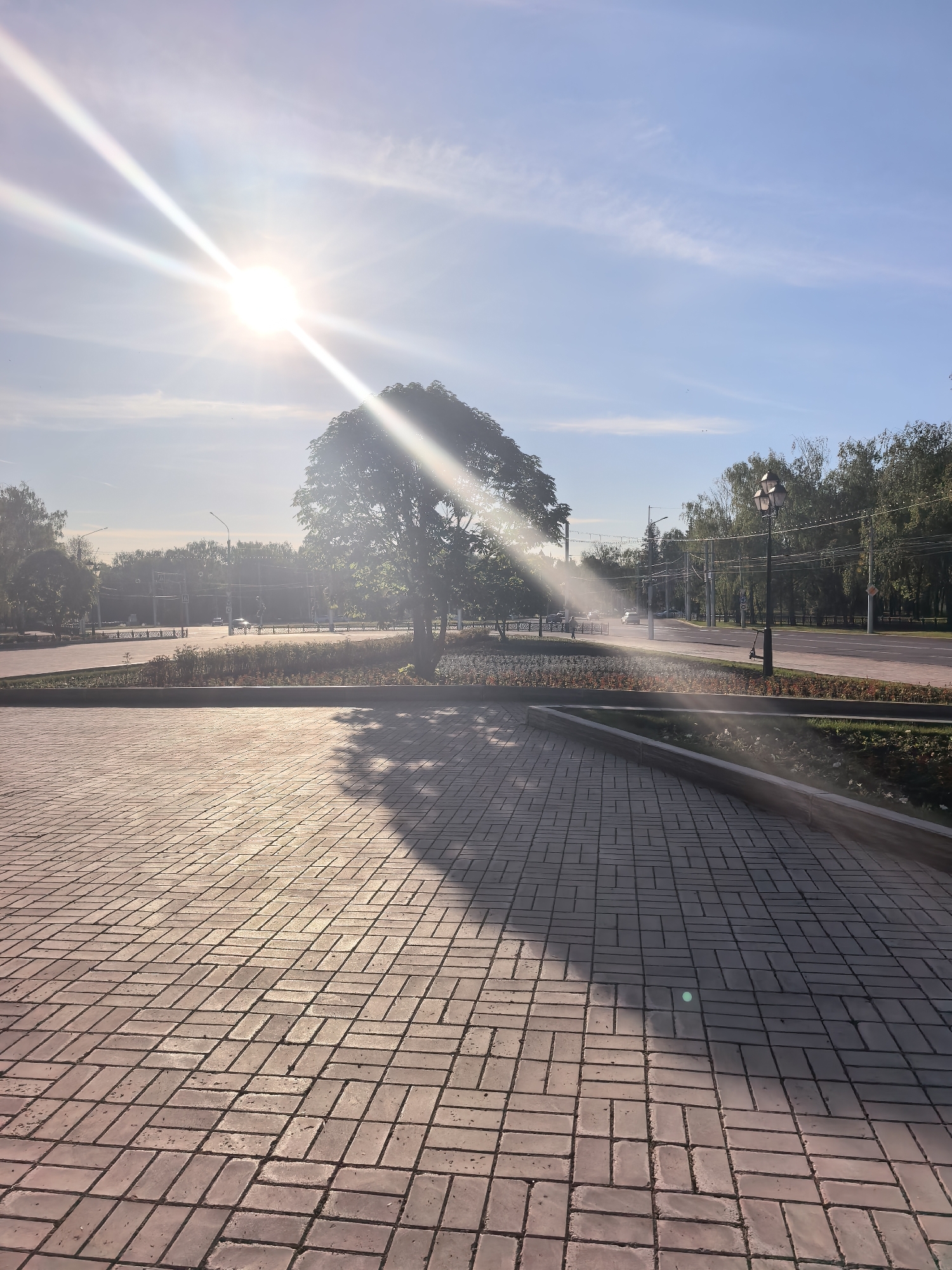 The image depicts a sun-drenched scene in Sterlitamak, Russia, at the intersection of Kommunisticheskaya and Khudayberdina streets. The foreground features a brick-paved plaza with the distinct shadow of a tree cast across it. Beyond the plaza is a strip of flower beds separating it from a four-lane road. On the far side of the road, trees line the street, interspersed with streetlights. Cars are visible on the road. The sun is high in the sky, emitting strong rays that partially obscure the scene. No people are immediately visible.