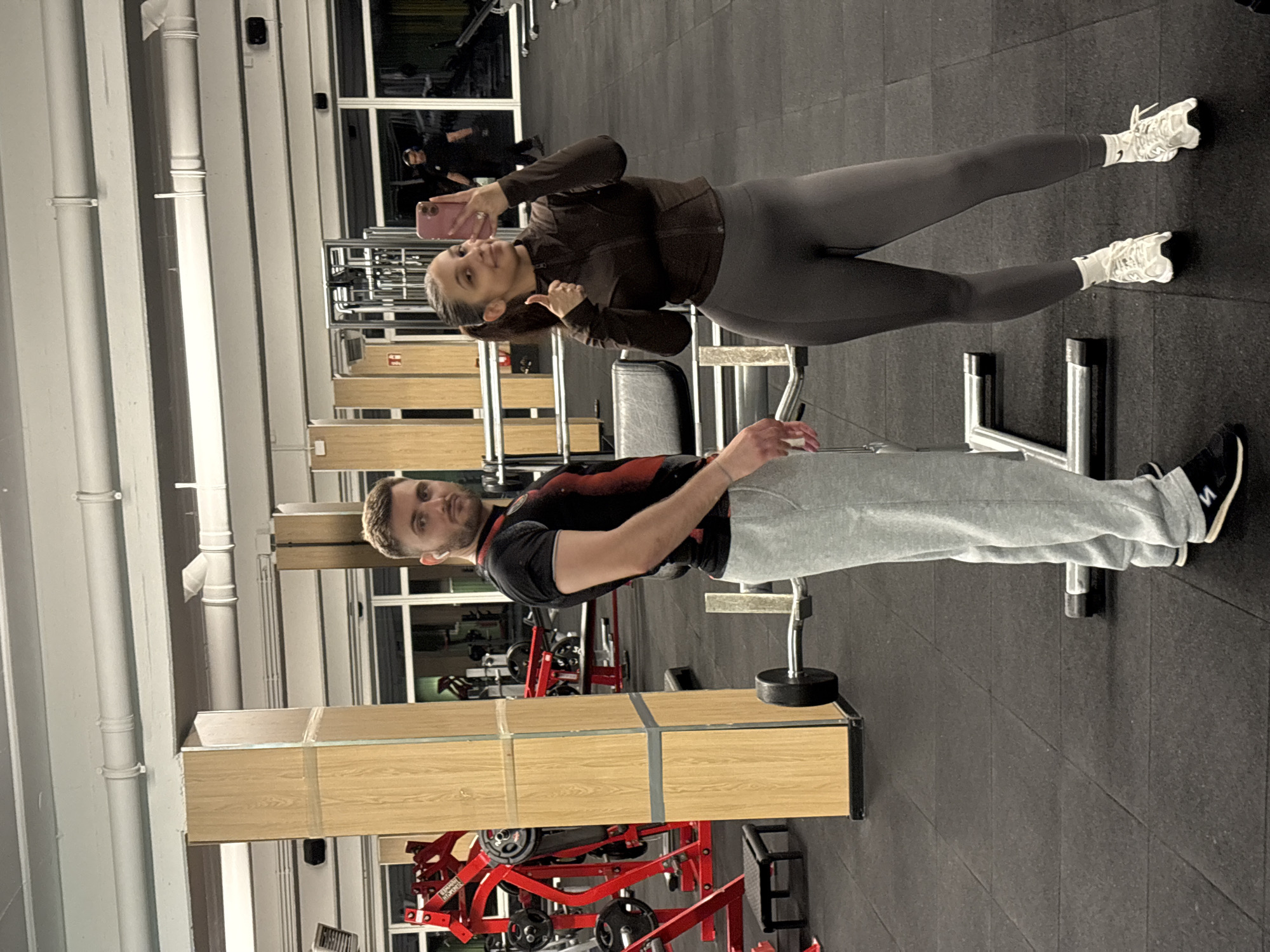 Inside a gym with black flooring, a woman in a brown jacket and grey leggings is holding a pink phone, taking a selfie. She is positioned above and slightly to the side of a man in a black and red shirt and grey sweatpants. Various pieces of gym equipment are visible in the background and foreground. The location is likely in Mönchengladbach, Germany.