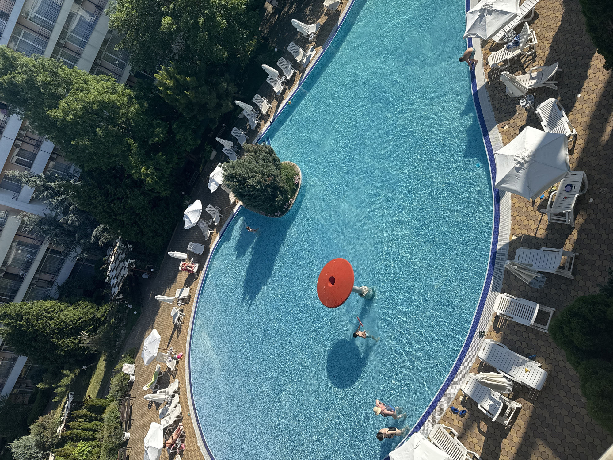A high-angle, full shot captures a bright, sunny day at a resort pool in Nesebar, Bulgaria. The foreground is dominated by the expansive, aquamarine water of the curved swimming pool, with dappled sunlight creating moving patterns on its surface. Several lounge chairs and white umbrellas are arranged along the paved poolside, some unoccupied, others with sunbathers.
In the pool, a few people are visible. Near the left edge, a woman in a black swimsuit and a woman wearing a wide-brimmed hat and a blue swimsuit are standing. Further into the pool, a child in blue shorts is playing with a red toy. Towards the center of the pool, a distinctive red mushroom-shaped water feature is present. In the distance, a lone figure is swimming or wading.
The background features lush green trees and the facade of a multi-story building, likely a hotel or resort complex, with balconies and windows visible. The poolside area is paved with hexagonal interlocking bricks. The overall scene depicts a relaxing vacation setting.
