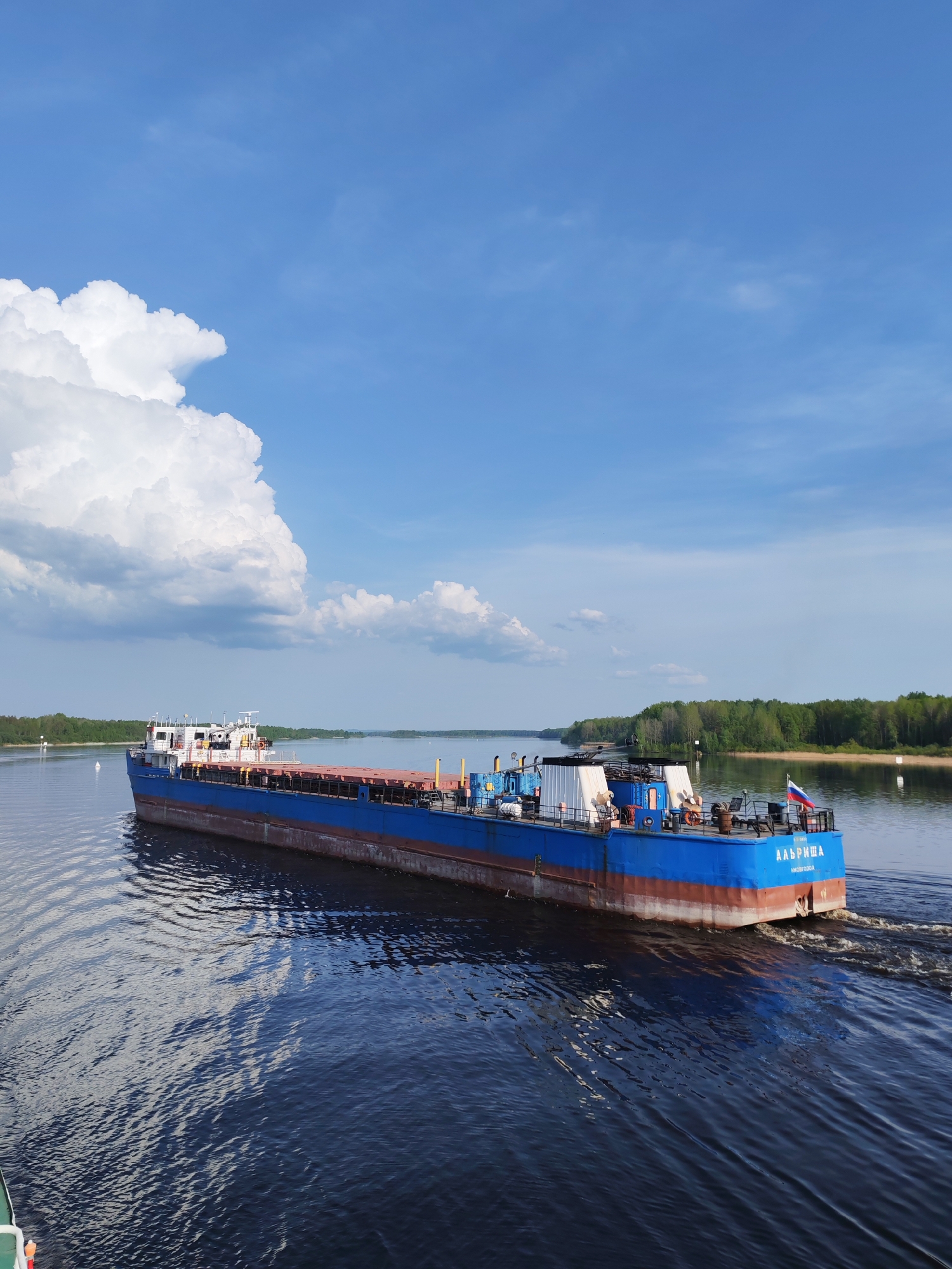 The image depicts a blue and red cargo ship named "ААБРИША" sailing on a body of water, likely a river or large lake. The ship has a Russian flag displayed at the stern. The surrounding environment features trees lining the shoreline and a blue sky with puffy white clouds. The location is Дурасово, Russia, as indicated by the ship's name "ААБРИША" and other factors. The water reflects the sky and clouds. No people are clearly visible in the image.