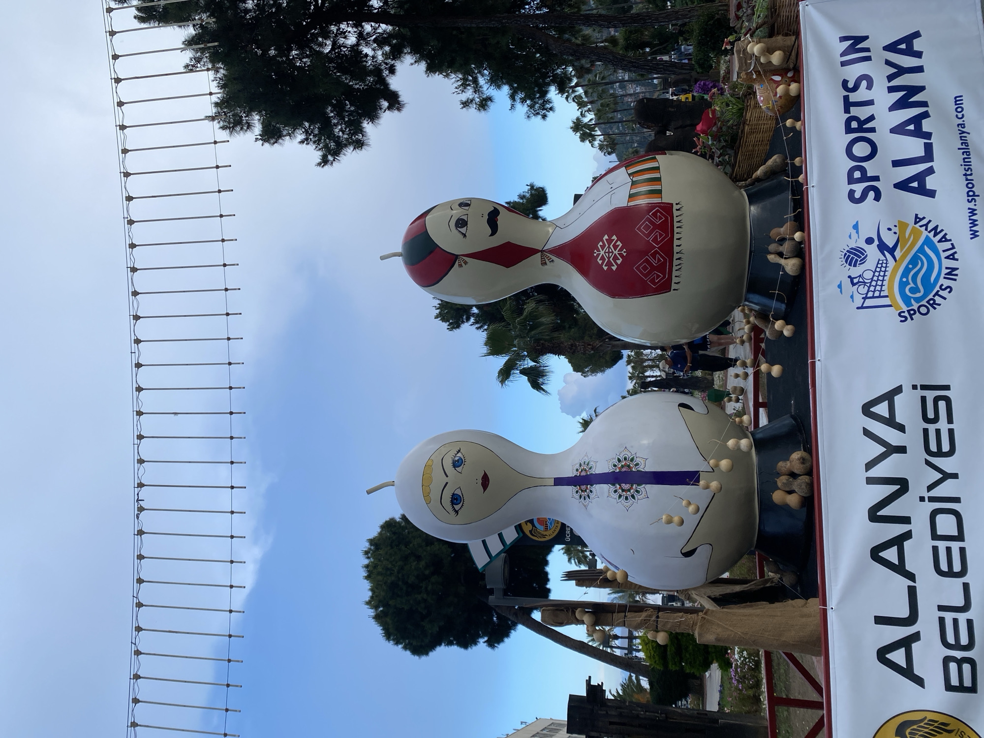 The image shows two large, gourd-shaped sculptures with painted faces and designs, situated in what appears to be a public space in Alanya, Turkey. One sculpture is painted with a male face, including a mustache, and red and white geometric patterns. The other is painted with a female face and purple designs. Both sculptures have strings of smaller gourds hanging around them. A banner to the right reads "SPORTS IN ALANYA" and "ALANYA BELEDIYESI." Trees and a section of a rope bridge are visible in the background. The sky is visible above the trees.