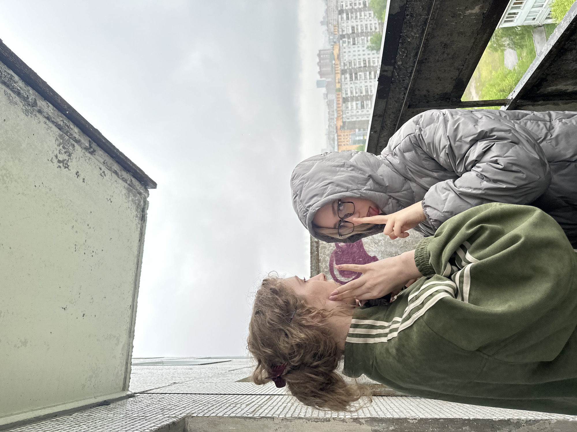 Two young women are on a rooftop overlooking a city. The woman closest to the camera wears a green sweatshirt with three white stripes on the sleeves and has curly brown hair. The other woman wears a grey puffy jacket with a hood, and glasses, and sticks her tongue out. They appear to be interacting playfully. They are standing on a metal grid surface. Part of a concrete wall is visible. The city in the background has mostly apartment buildings and an overcast sky. The location is likely Krasnoyarsk, Russia.