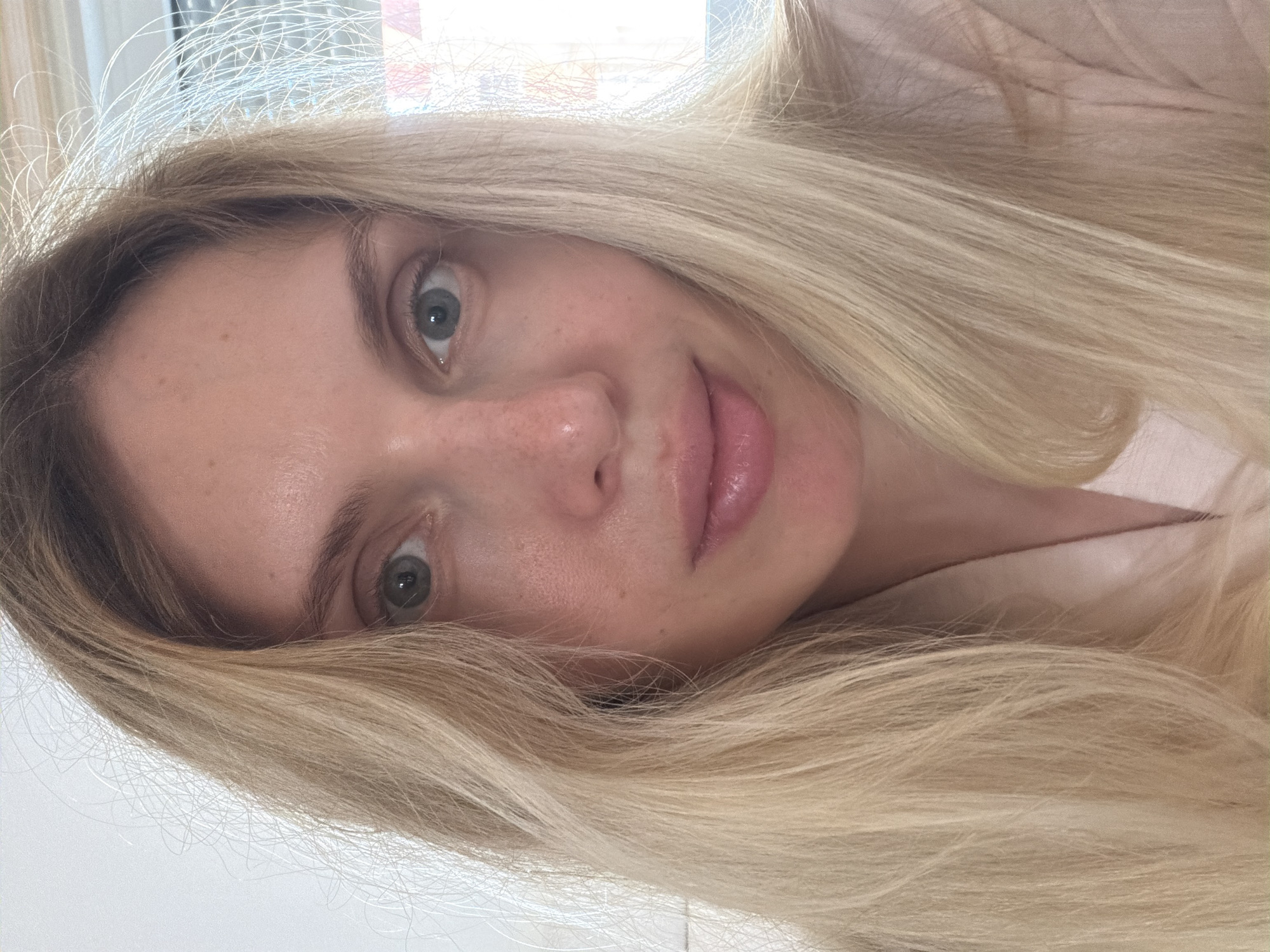 A close-up, slightly blurred selfie shows a woman with long, blonde hair. The woman has fair skin and light-colored eyes. Some freckles are visible on her nose. The background appears to be a bright, indeterminate indoor space. There is no evidence to pinpoint the location to Budva, Montenegro.