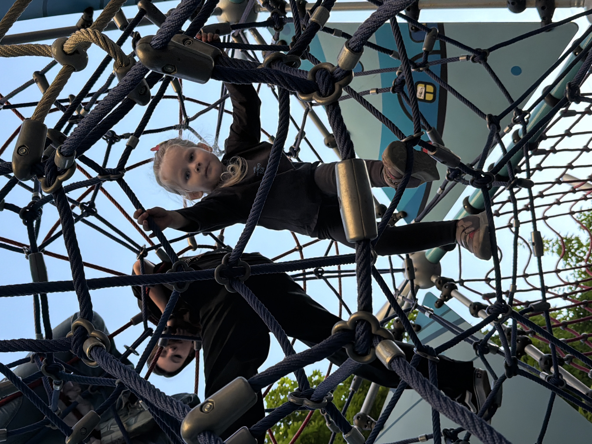 The image depicts a scene at a playground in Khimki, Russia, featuring two individuals engaged with a large, intricate rope climbing net. The primary subject is a young girl with blonde hair, pulled back with a pink clip, wearing a dark long-sleeved shirt, dark pants, and light-colored sneakers with subtle pink details. She is positioned within the upper section of the rope net, looking directly at the camera with a neutral expression. Her hands are gripping the ropes, and her body is partially obscured by the dense network.
Below and behind her, a second person, likely an adult or older child, is also visible on the climbing structure. Their head is seen looking downwards, and they are wearing dark clothing, largely integrated into the ropes.
The climbing net itself is a prominent feature, constructed from thick, dark blue/black and lighter tan/grey ropes, connected by various metal or robust plastic fasteners. This creates a complex, three-dimensional mesh. In the background, a clear, bright blue sky suggests a daytime setting with fair weather. Parts of a light blue and white painted structure, possibly a platform or panel of the playground, are visible through the ropes. Some green foliage, likely from distant trees or bushes, can be seen in the lower right and through parts of the net. No discernible text is visible on any of the subjects or objects. The scene conveys an atmosphere of active outdoor play.