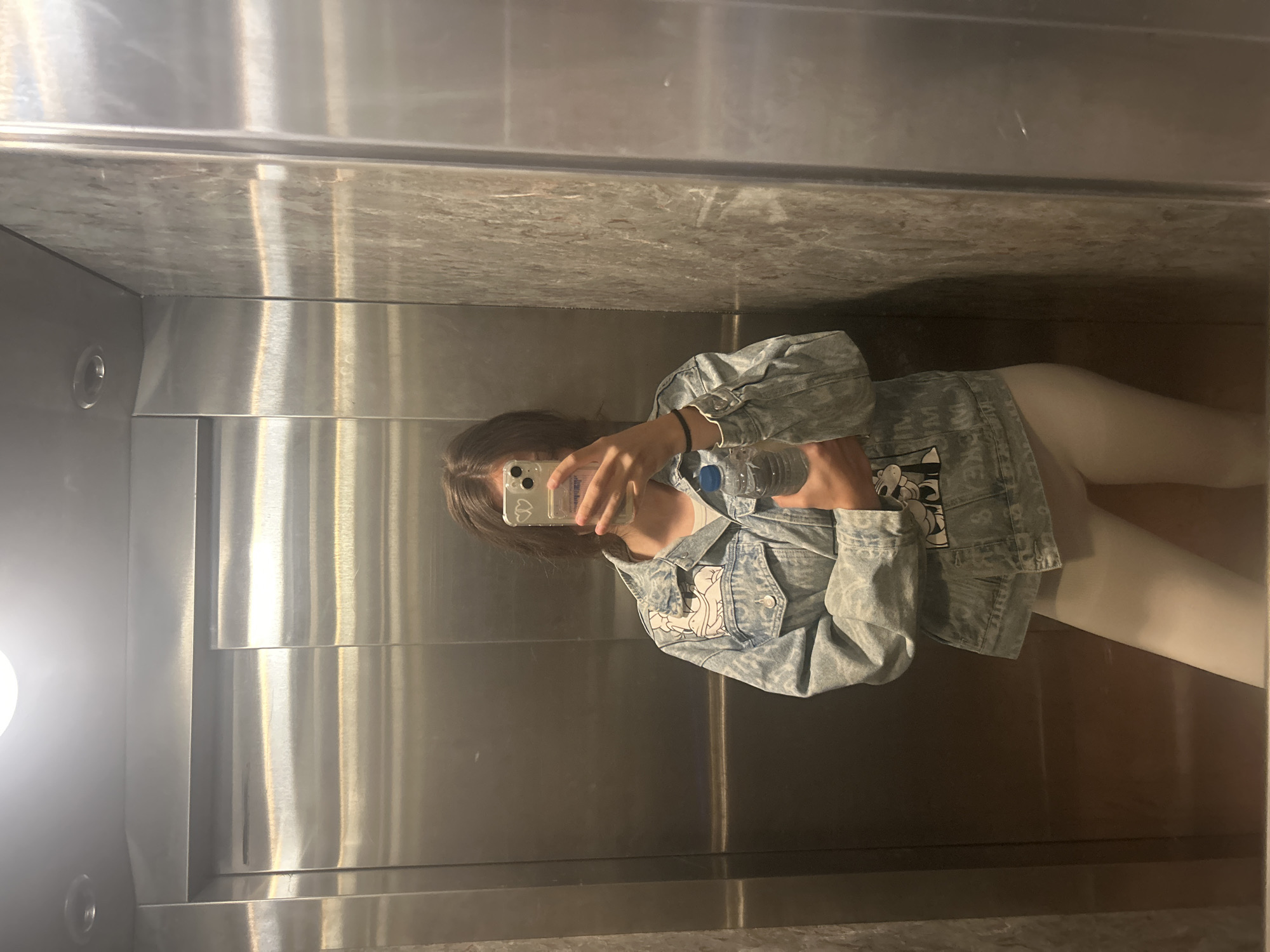 A woman stands in an elevator taking a selfie. The elevator has stainless steel walls and a marble-look ceiling. The woman wears a light blue denim jacket and white leggings. She is holding a water bottle and a cell phone. There is no indication in the image that this is in Burgas, Bulgaria.