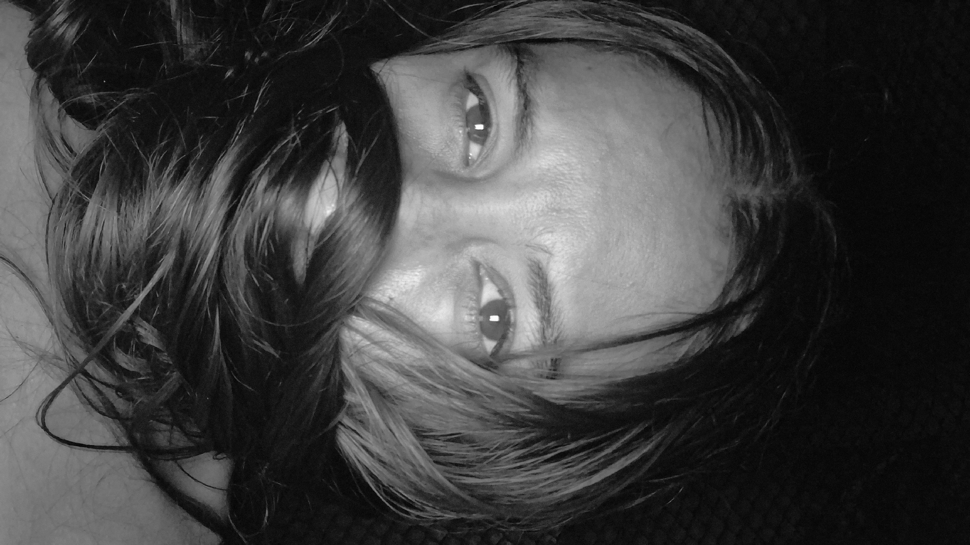 A black and white close-up photograph shows the upper half of a person's face, with their hair obscuring their mouth and nose. The person's eyes are visible and looking directly at the camera. The lighting is dim, with the background being a dark, textured surface. The image appears to be a selfie, taken indoors. The individual's skin shows some texture and natural light reflections. The hair is dark and appears to be wet or oily, with strands falling across the face. There is no information about the location in Vallecito, Venezuela.