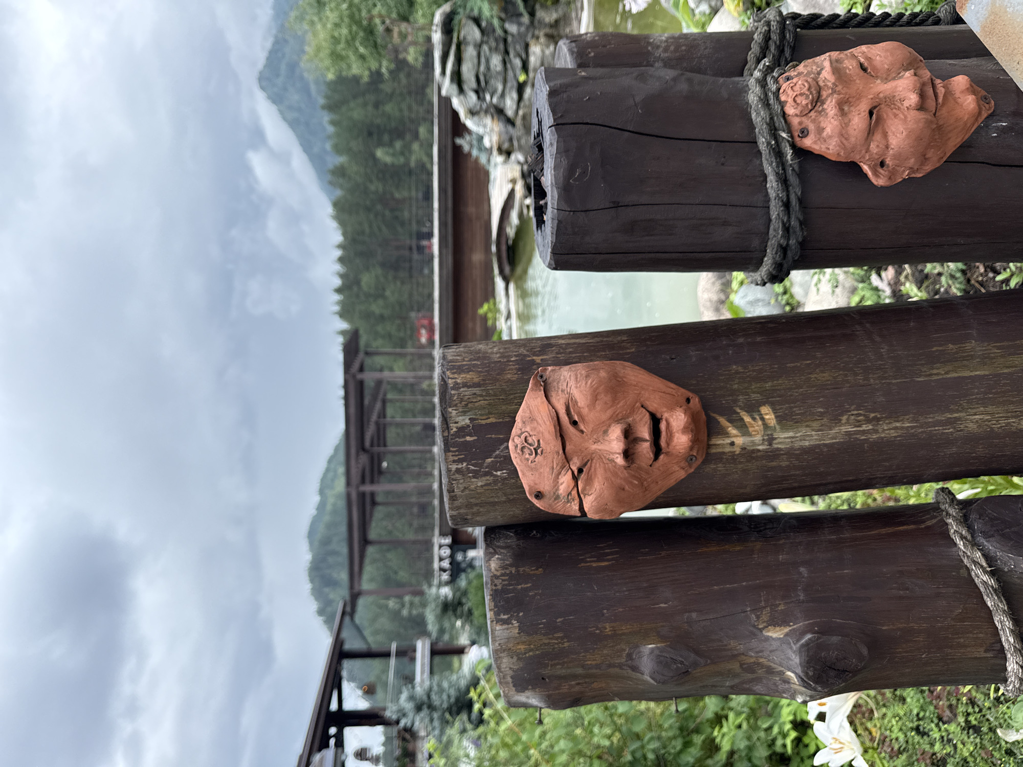 In Barangol, Russia, two rustic wooden posts stand in the foreground, each adorned with an orange clay mask resembling a face. The mask on the left post is larger and shows a cracked face with closed eyes and an open mouth. The mask on the right post is smaller and features closed eyes and a slightly open mouth. The posts are bound with thick rope. Behind the posts, a body of water reflects the overcast sky, with a small arched wooden bridge spanning a section of it. In the background, a dense forest of green trees lines the horizon, with hazy mountains rising beyond. Structures, possibly part of a resort or cafe, are visible in the mid-ground, with a sign reading "КАФЕ" (Cafe) partially obscured. The overall atmosphere is one of a natural, somewhat weathered outdoor setting.