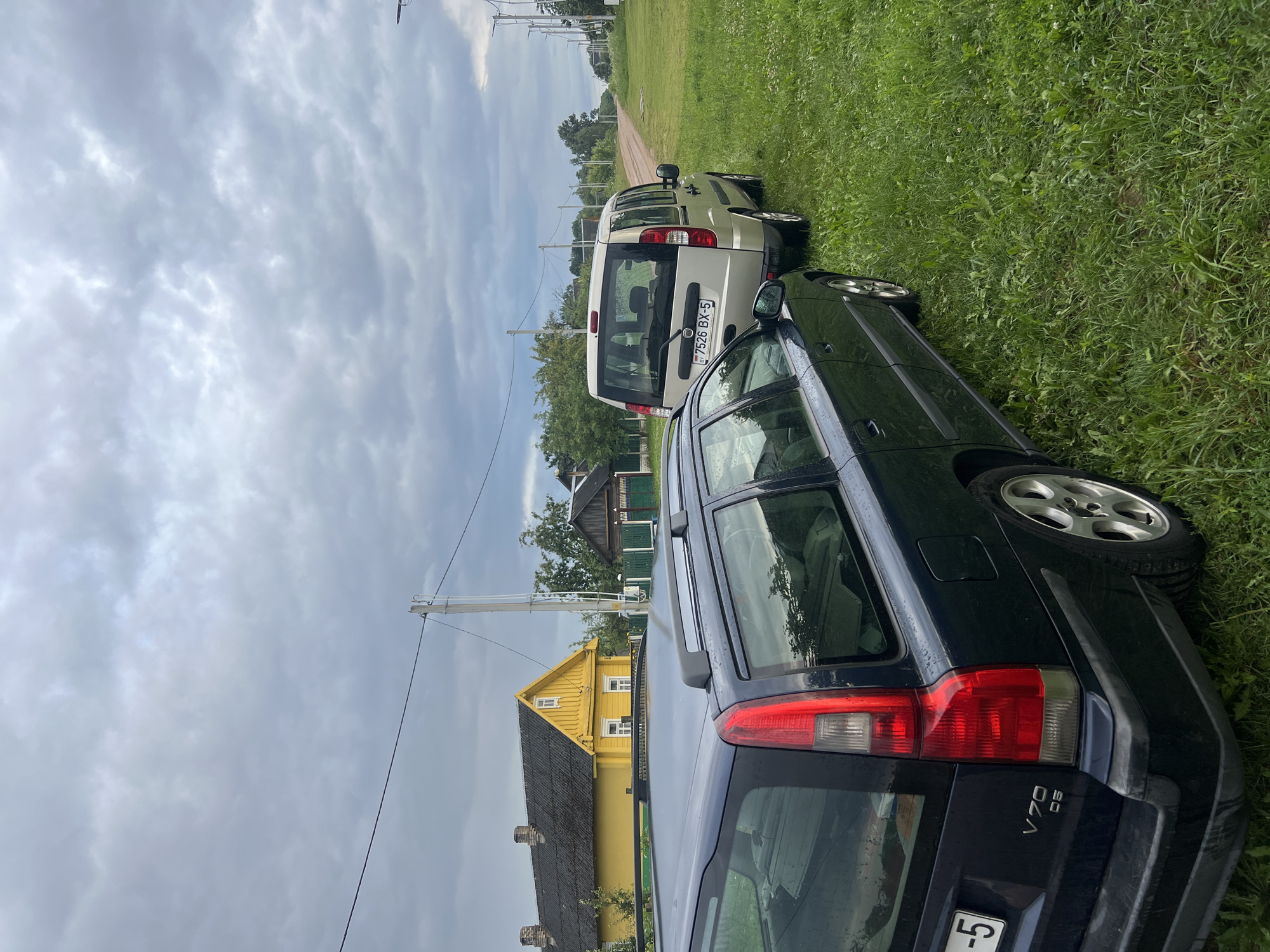 The image captures a rural scene in Бучацінскі сельскі Савет, Belarus. In the foreground, a dark blue Volvo V70 station wagon is parked on the left side of a gravel road. The car is facing away from the camera, with its rear visible. To the right of the Volvo, a silver minivan, possibly a Fiat Doblo, is also parked on the grass verge, facing the same direction. The minivan's license plate reads "7526 BX-5 BY".
A bright yellow house with a dark roof and white trim is situated behind the Volvo on the left side of the road. Further down the road, more houses and trees can be seen. The sky is overcast with grey clouds, suggesting a potentially gloomy or rainy day. The road itself appears to be unpaved, made of gravel or dirt, and it curves to the right as it recedes into the distance. Utility poles with wires are visible along the side of the road. The area is covered in lush green grass, indicating a rural or semi-rural setting.