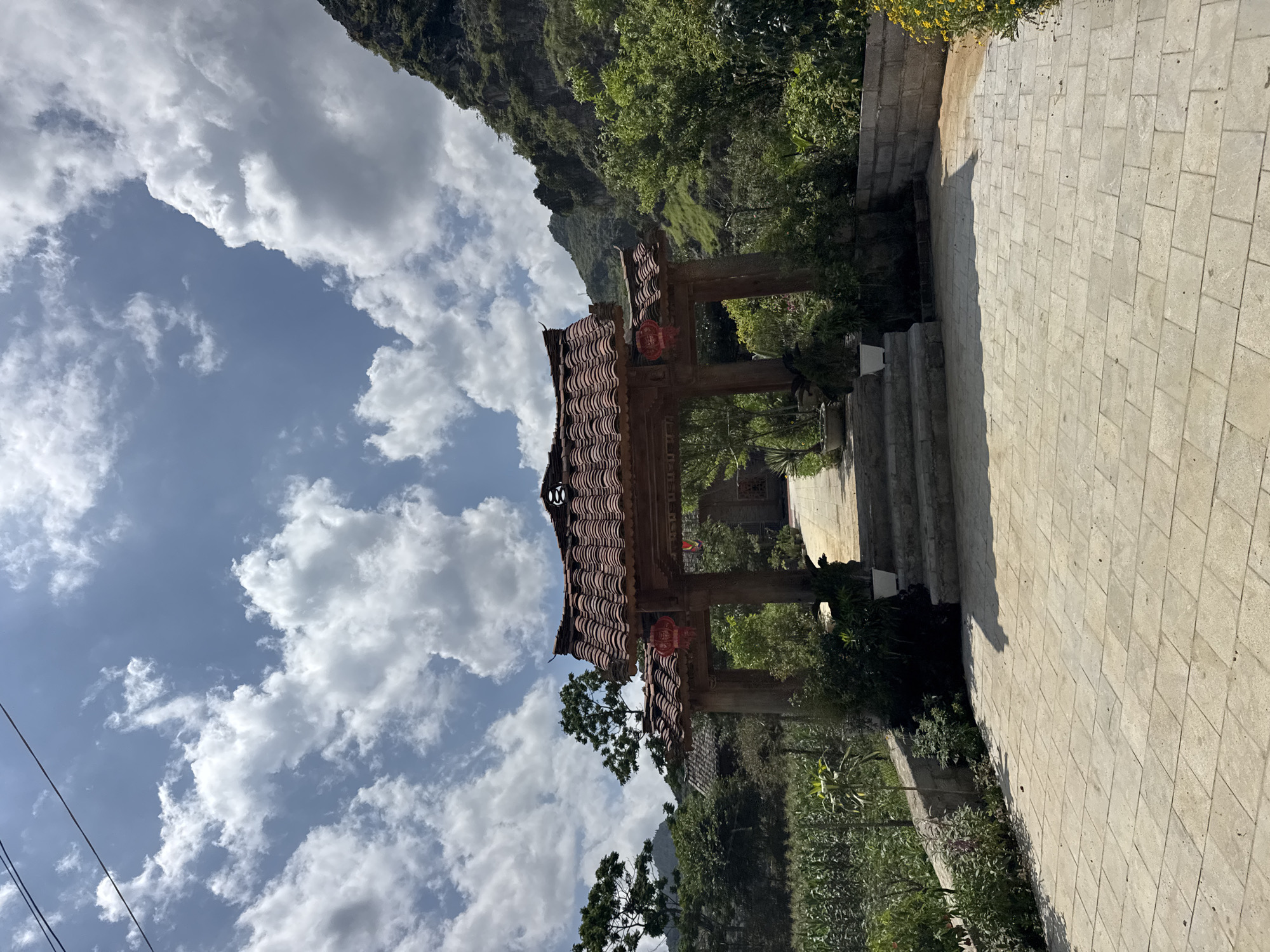 The image captures a scene in Đồng Văn Commune, Vietnam. A stone-paved path, slightly elevated with steps, leads towards a traditional, wooden Asian gate structure with a tiled roof. The gate is adorned with red lanterns. The path and gate are flanked by greenery. Beyond the gate, the landscape includes a lush, mountainous terrain. The sky above is partially covered with white, puffy clouds against a blue backdrop. A power line is visible on the left side of the image.