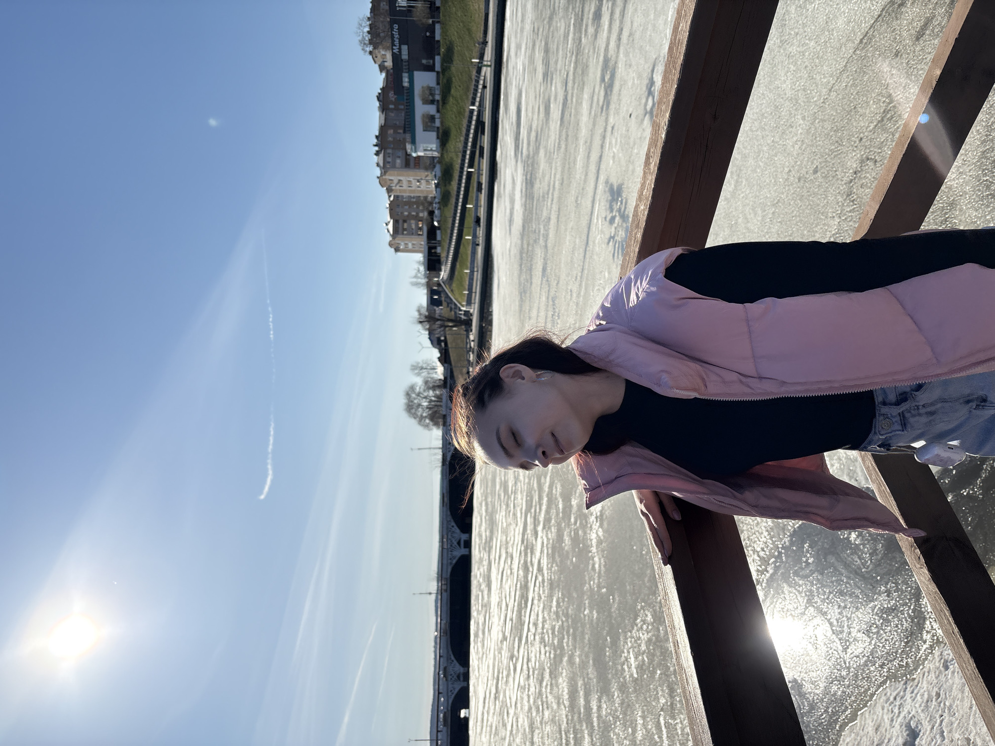 A young woman is captured outdoors, leaning against a wooden railing next to a large body of water, possibly a river or canal, under a bright, clear blue sky. She has dark hair and is wearing a black long-sleeved top, a light pink sleeveless puffer vest, and light-wash blue jeans. Her head is tilted downwards, and her eyes appear closed or cast down, suggesting a moment of contemplation or rest.
The water surface is highly reflective and appears partially frozen or covered in ice, strongly reflecting the dazzling sunlight. The sun itself is visible in the upper left of the sky, creating a radiant glare. In the background, across the water, a cityscape unfolds with a mix of buildings, some with multi-story European architectural elements, and areas of greenery with trees. A distinctive bridge with intricate railings can be seen further down the waterway.
The weather is sunny and clear, indicated by the bright sky with only a few faint contrails. The woman's puffer vest and the icy appearance of the water suggest it might be a cool or cold day. A sign displaying the text "Maestro" is visible on one of the distant buildings. The scene is set in Votkinsk, Russia.