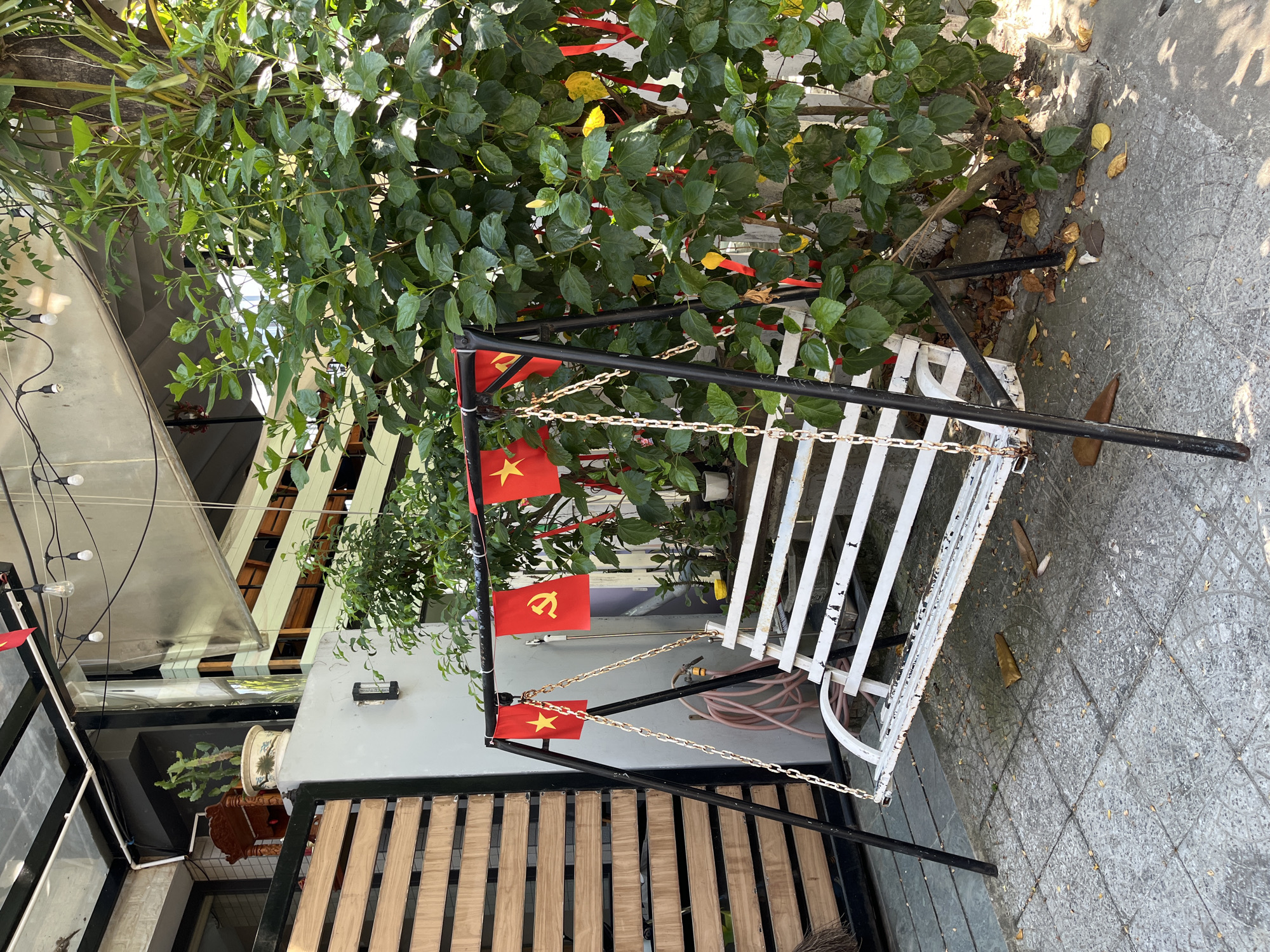 The image depicts a white, metal swing set with black supports, adorned with small Vietnamese flags, positioned on a tiled surface next to a building and under a leafy green shrub. The building features a modern design with wood slats and a white exterior. The scene is located outdoors on a paved area. The presence of Vietnamese flags and the architectural style suggest the location is likely in Vietnam, possibly in a public space or a business establishment. The precise location within Đà Nẵng cannot be determined from the image.