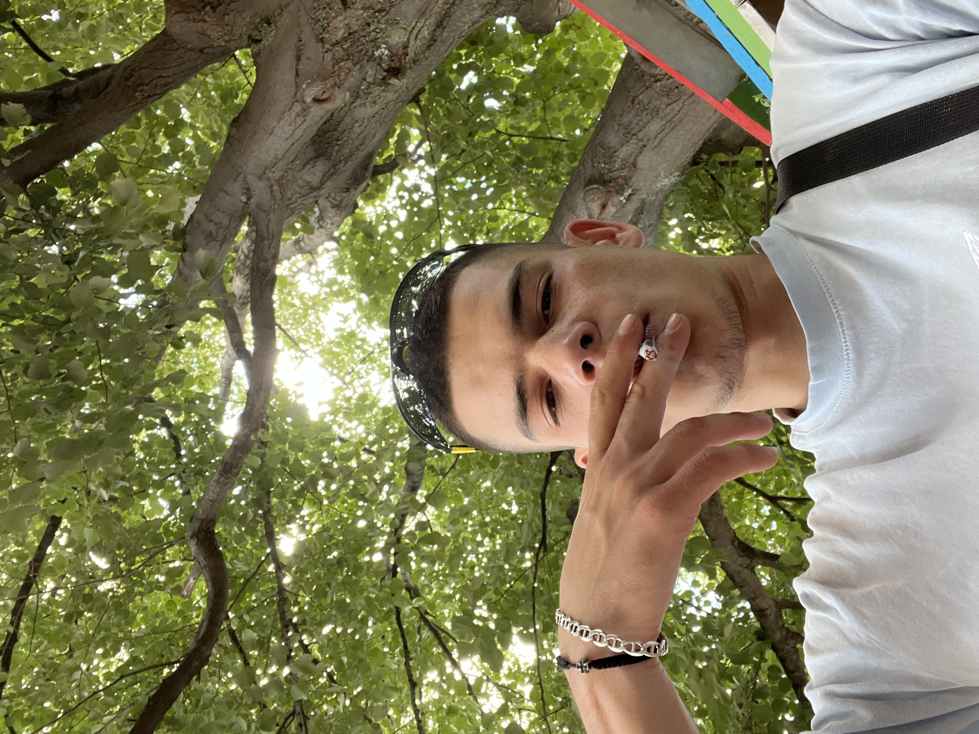 A young man with short dark hair and dark eyes is holding a cigarette to his lips, exhaling smoke. He is wearing a light blue t-shirt and sunglasses pushed up onto his head. He has a silver chain bracelet on his left wrist. He is outdoors, with a large tree providing shade from above, dappled sunlight filtering through the leaves. The background shows the trunk of a large tree and a section of what appears to be a colorful structure. The image is a low-angle, selfie-style shot. The man's location is the city of Sopot, Bulgaria.