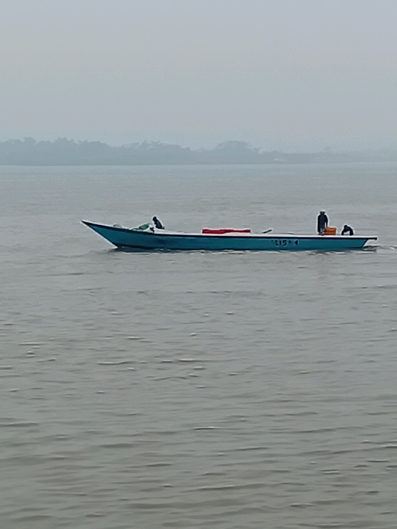 A long, narrow, light blue boat with a pointed bow is traveling across a body of calm, grey water. The boat has a dark stripe along its side with the white numbers "LIS 4" painted on it. Two individuals are visible on the boat; one is standing near the stern, facing away from the camera, and appears to be wearing dark clothing. Another person is crouched or sitting near the bow. A red object is positioned on the deck towards the bow, and a small orange container is located near the standing figure. The background shows a hazy, treeline of dense foliage with a pale, overcast sky. The overall atmosphere is muted and diffused by mist or fog. The location is Sungai Meriam, Indonesia.
