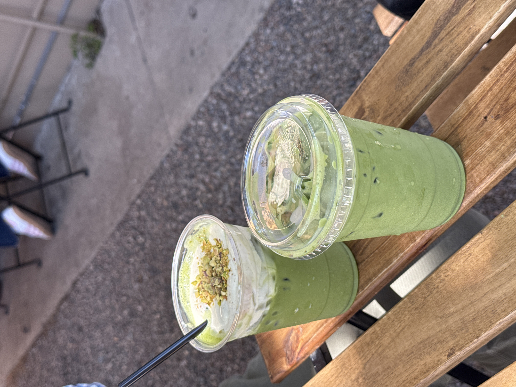 Two plastic cups of green matcha drinks are on a wooden outdoor table in Chula Vista, United States of America. The cup on the right has ice and a clear plastic lid. The other cup has a black straw and whipped topping covered with chopped nuts.