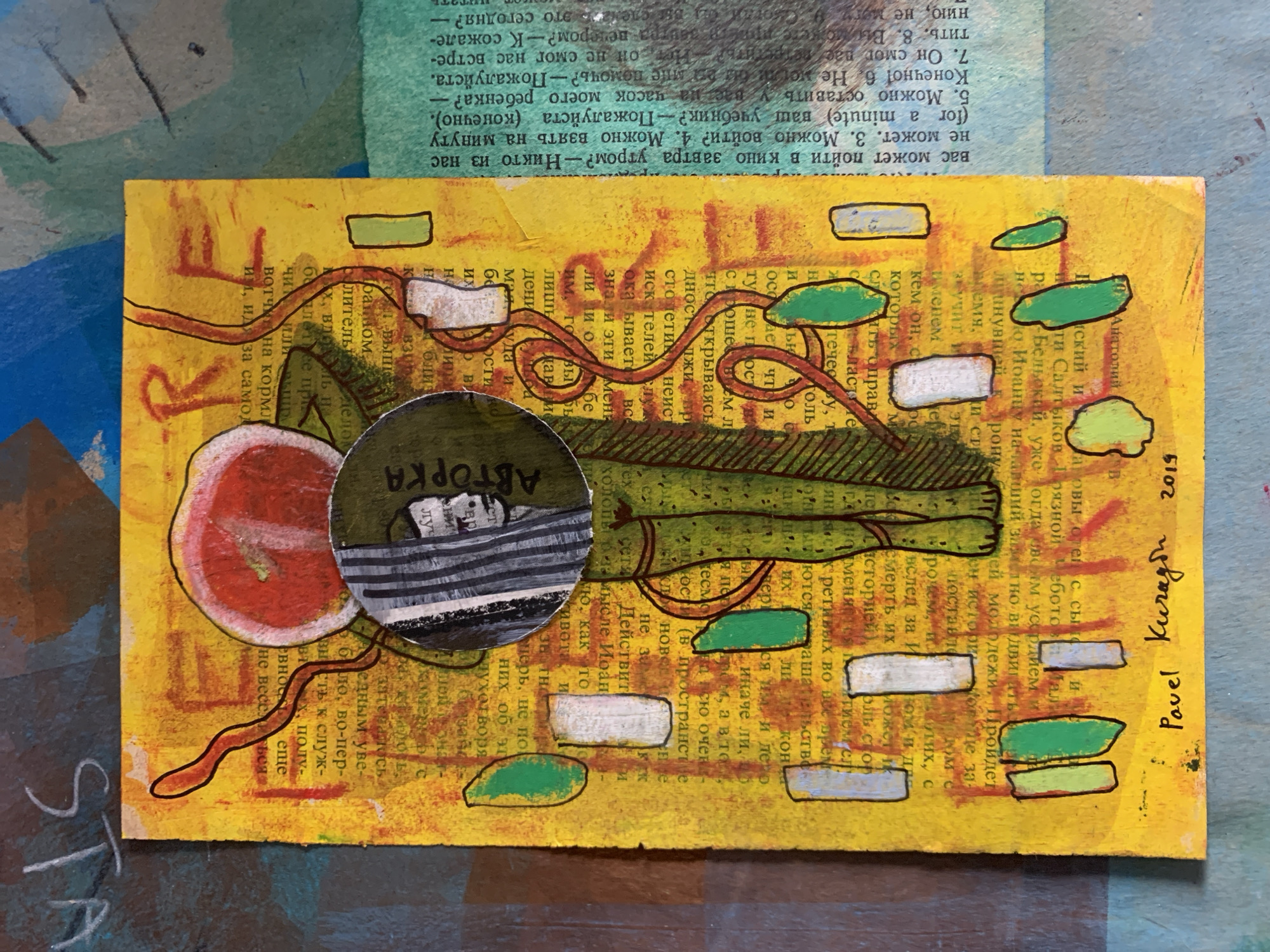 The image is a close-up, high-angle shot of a mixed-media artwork on a yellow, textured surface, likely paper, that resembles aged book pages. The artwork is a collage and painting that depicts a stylized, green figure with red, vine-like elements extending from its head. The figure's head is covered by a circular element with vertical black and white stripes, and a smaller, black and white image of a person's face is visible within this circle. The text on the yellow background is in Russian and appears to be from a book. There are several rectangular shapes, some painted green and white, scattered across the surface. In the bottom right corner of the artwork, the signature "Pavel Kuragin 2019" is visible.
The artwork itself is not located in Sergeevo, Russia. There is no visual information in the image that connects it to Sergeevo or any other specific city. The image solely displays a piece of art.