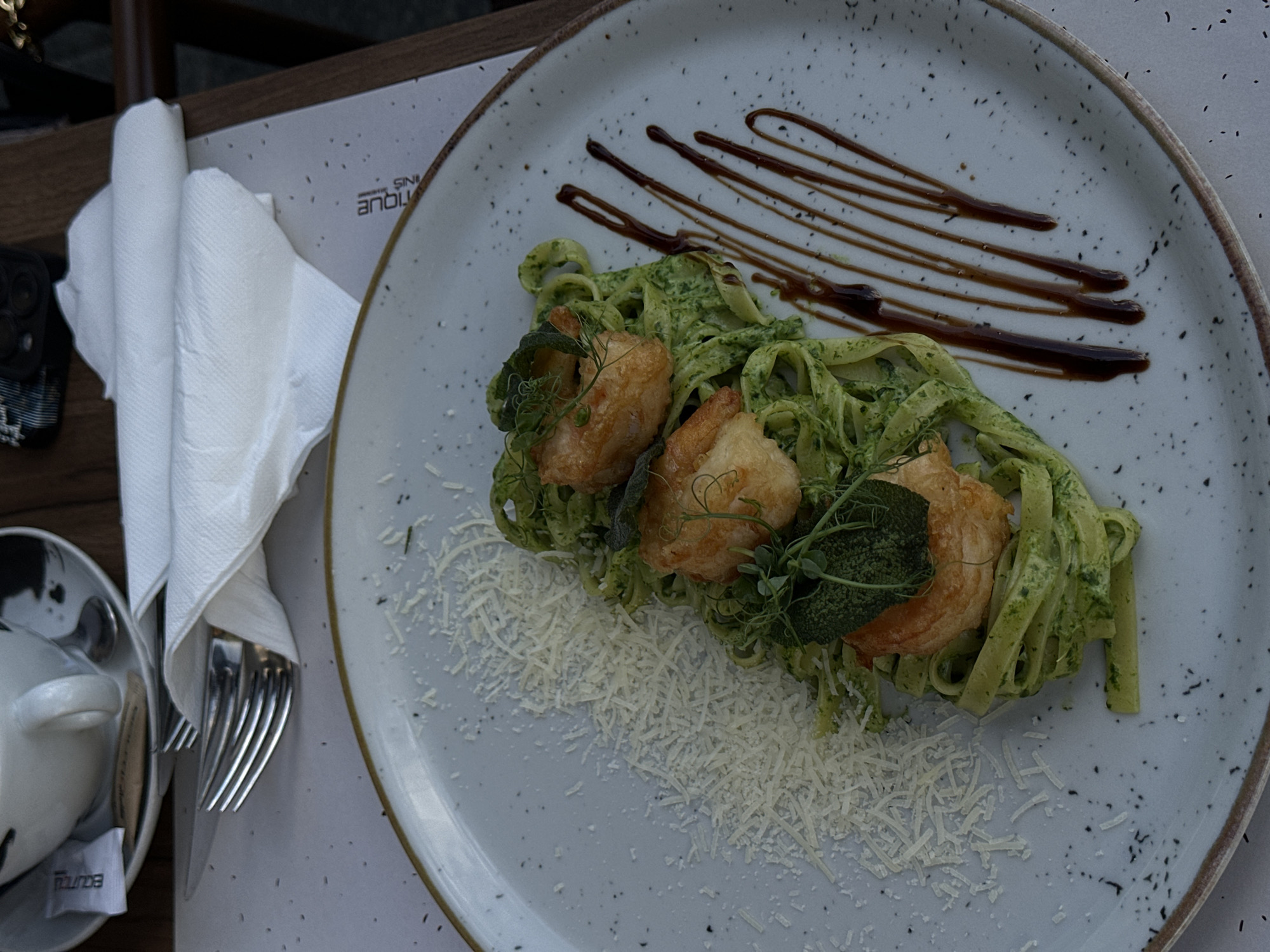 The image displays a close-up, elevated view of a meticulously presented meal on a table, likely in a restaurant setting in Belgrade, Serbia. The central focus is a light grey ceramic plate with dark speckles and a brown rim, holding a serving of pasta with shrimp.
The pasta, appearing to be fettuccine or tagliatelle, is coated in a vibrant green, creamy sauce, possibly pesto or a herb-based sauce. Three large, golden-brown shrimp are arranged on top of the pasta, garnished with fresh green microgreens (like pea shoots) and a couple of broader green leaves, possibly sage. A generous amount of grated white cheese, resembling Parmesan, is scattered along the lower left edge of the plate. The upper right portion of the plate is adorned with several artistic drizzles of a dark, glossy sauce, likely balsamic glaze.
To the left of the plate, a neatly folded white napkin partially covers a set of cutlery, including a silver fork and knife. Below the napkin, part of a dark mobile phone with visible camera lenses is resting on the table. Further to the left, a small, white ceramic cup or bowl, possibly for coffee or a condiment, is visible, containing a silver spoon and a white sugar packet. The table surface appears to be made of dark wood.
Visible text includes an inverted word, likely part of a brand or restaurant name, on a paper mat beneath the napkin, which reads "ANTIQUE" or similar (appearing as "ANBITIQUE" with other letters like "SINI"). On the sugar packet in the cup, partially obscured and inverted, the word "EOUUBA" is visible, likely "BAQUBA" or a similar brand name. The lighting is somewhat subdued, suggesting an indoor evening ambiance or a shaded outdoor area.